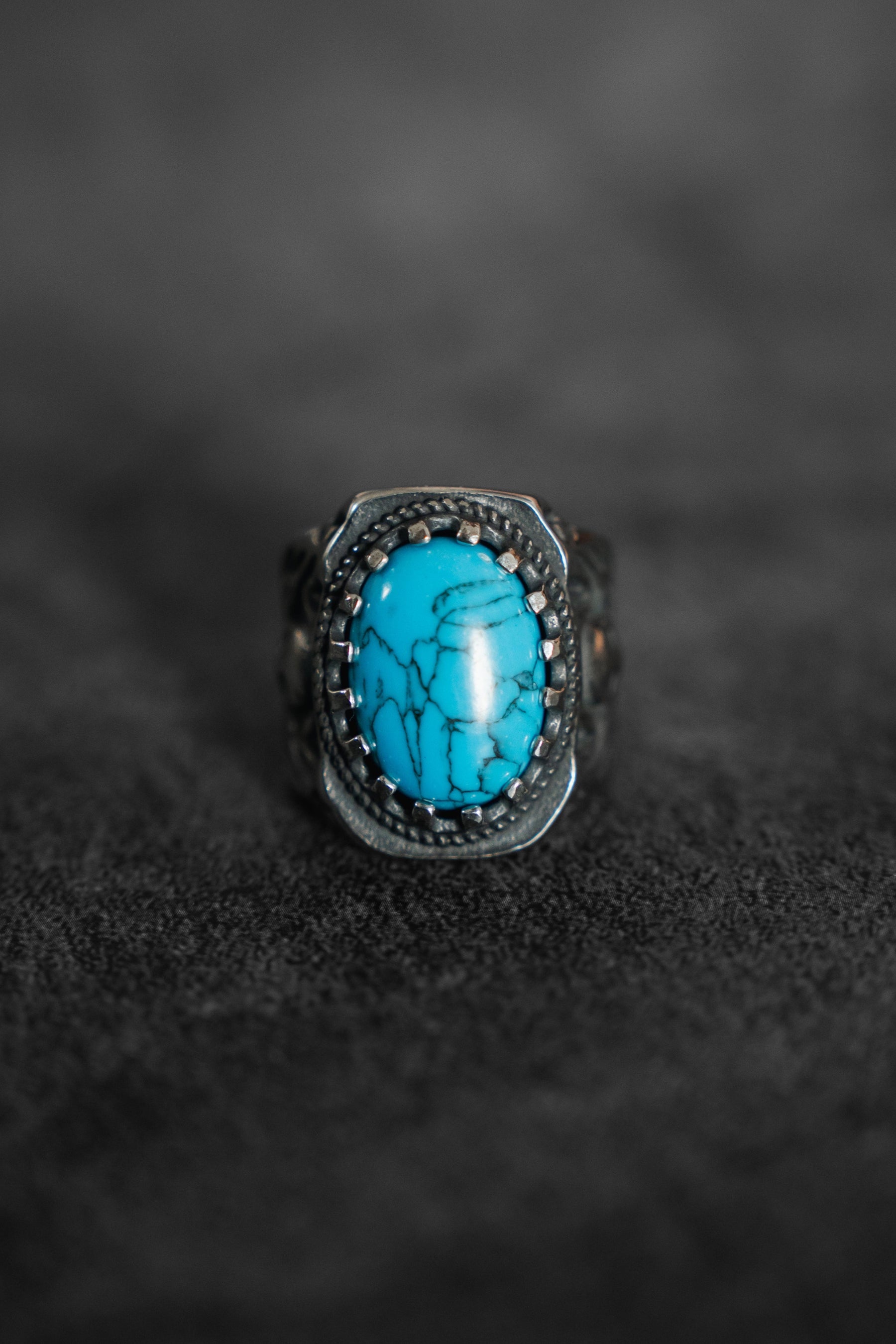 The Outsider Ring by Relentless Betrayal features a silver band with an ornate design and a marbled oval turquoise stone, set against a dark, textured background.