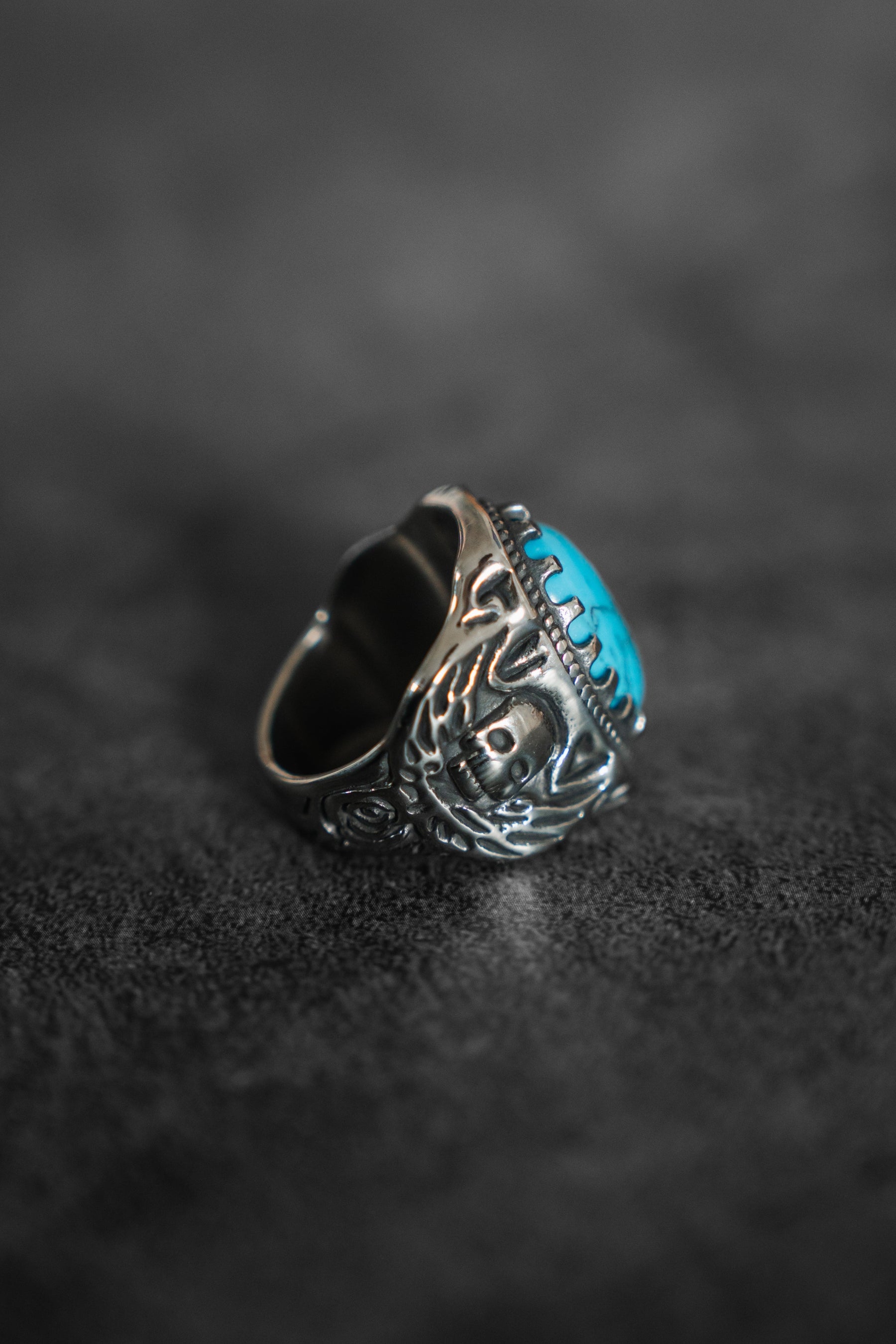 The Outsider Ring by Relentless Betrayal is a handcrafted silver piece featuring intricate designs and a large, oval turquoise stone. It sits elegantly on a textured dark surface, highlighting its unique charm.