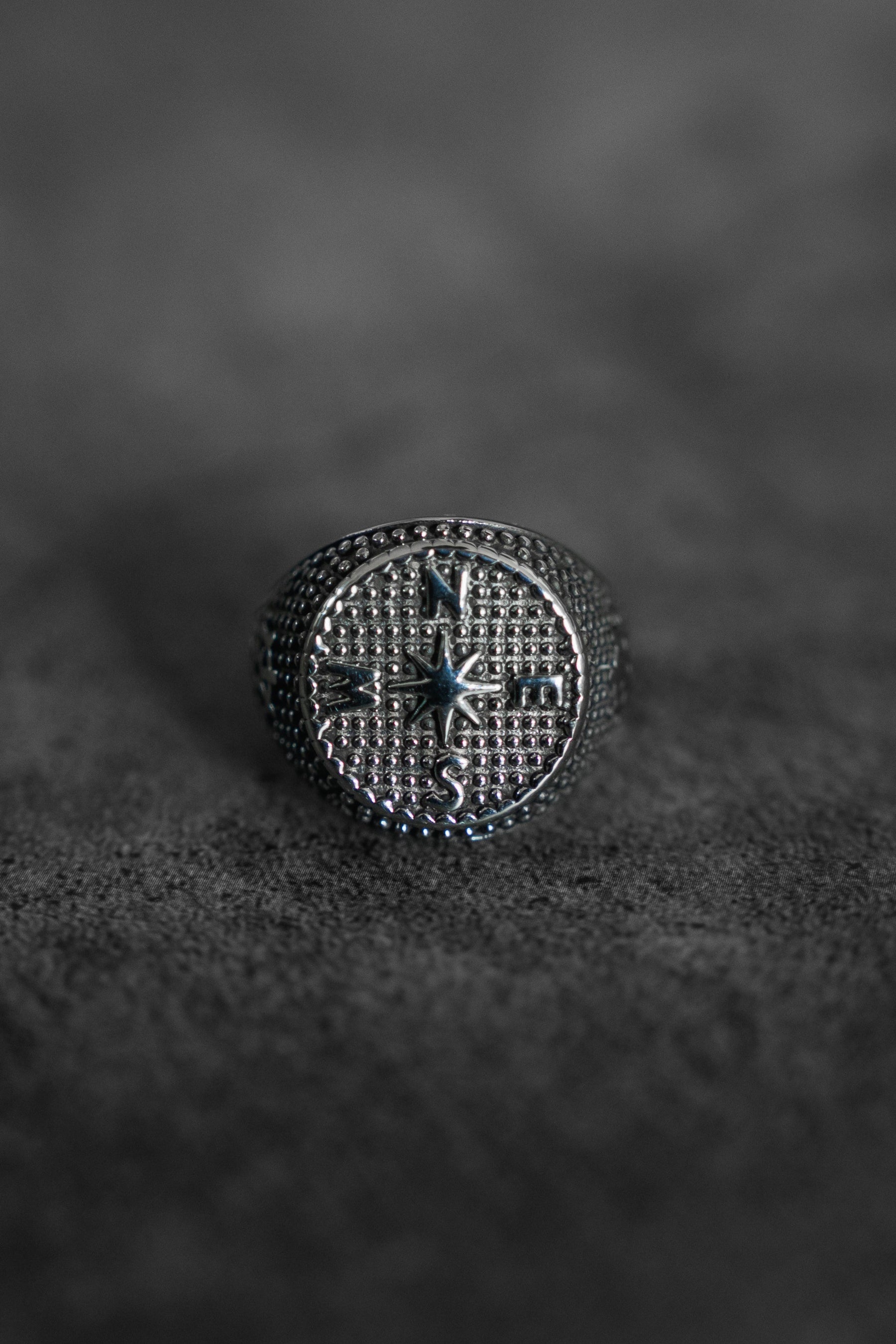 A close-up showcases the handcrafted Crossroads Ring by Relentless Betrayal in stunning silver, featuring a compass design with a central star and cardinal directions. This exquisite ring elegantly rests on a textured gray surface.