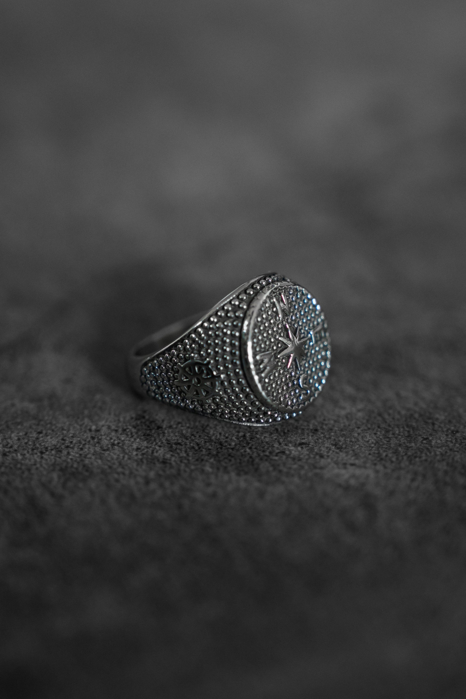 A close-up of the handcrafted Crossroads Ring from Relentless Betrayal on a dark, rough surface. The stainless steel ring showcases a central star design with raised dots and intricate patterns on the band, set against a softly blurred background.