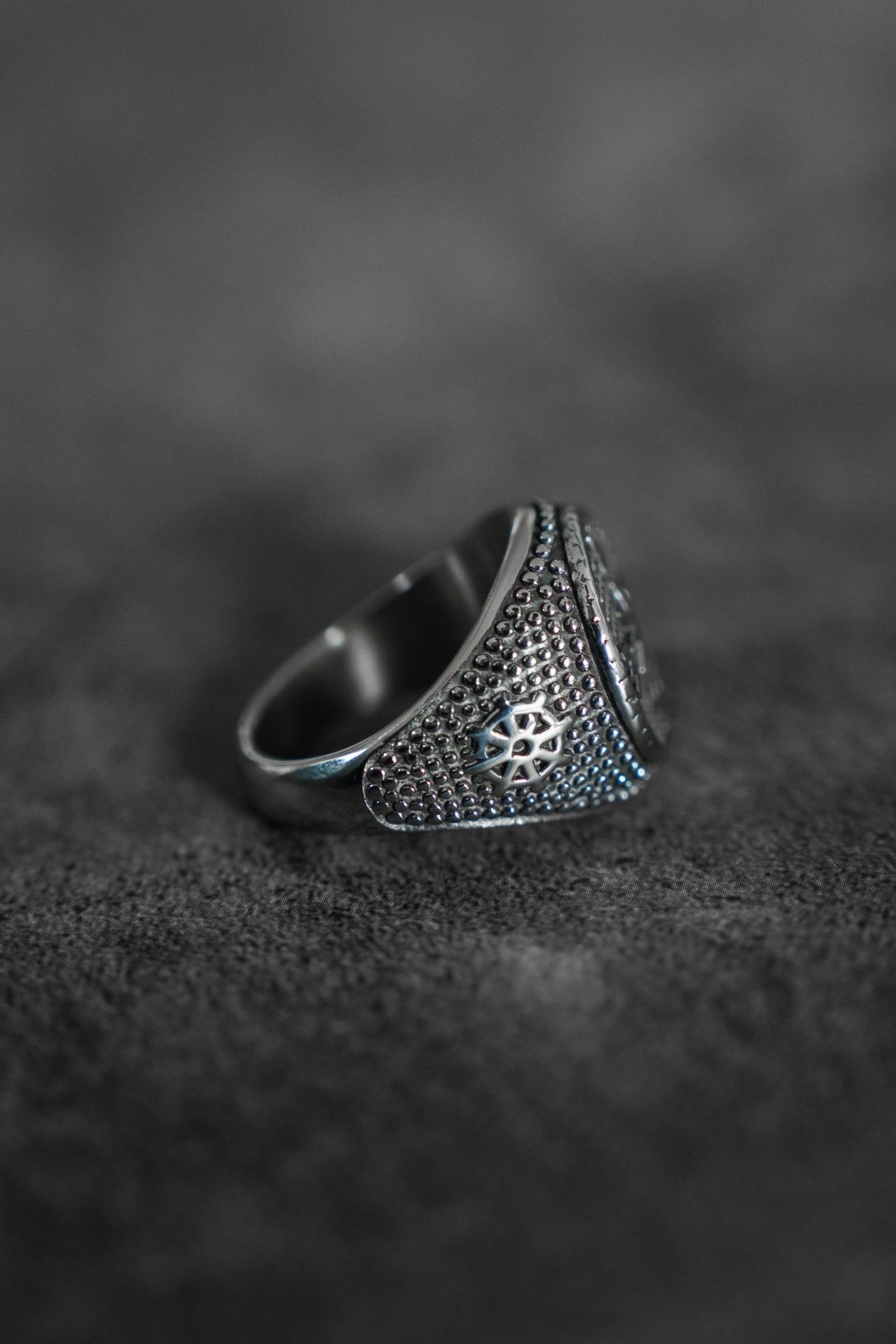 The Crossroads Ring by Relentless Betrayal, a handcrafted silver ring with intricate detailing, rests on a dark, textured surface. Its wide band showcases a raised geometric design that highlights its ornate craftsmanship.