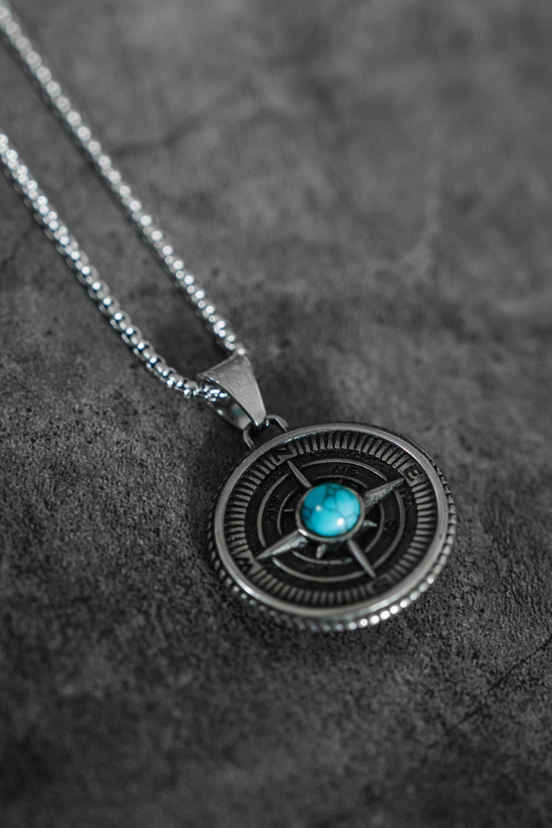 The Crossroads Necklace by Relentless Betrayal is a stunning silver piece with a round pendant, detailed designs, and a central turquoise stone symbolizing transition. Displayed on textured gray, it captures the essence of pivotal moments.