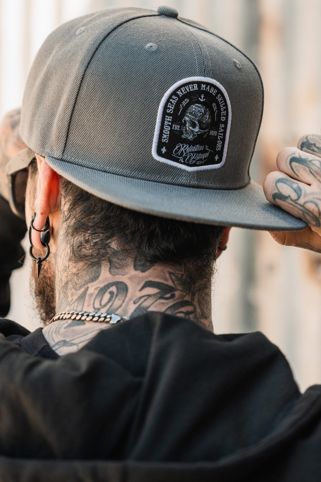 A person with tattoos on their neck adjusts a Relentless Betrayal Skilled Sailor Snapback, gray with a Smooth Seas Never Made Skilled Sailors patch. They wear a black jacket and earrings, facing away from the camera.
