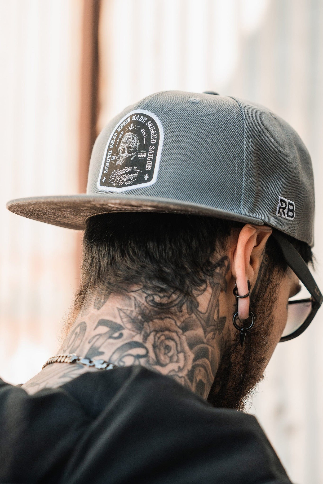 A tattooed individual with ear piercings is wearing the gray Skilled Sailor Snapback by Relentless Betrayal, made from a polyester cotton blend. The cap features a decorative patch and RB initials on the side. They have glasses and a short beard, facing away from the camera.
