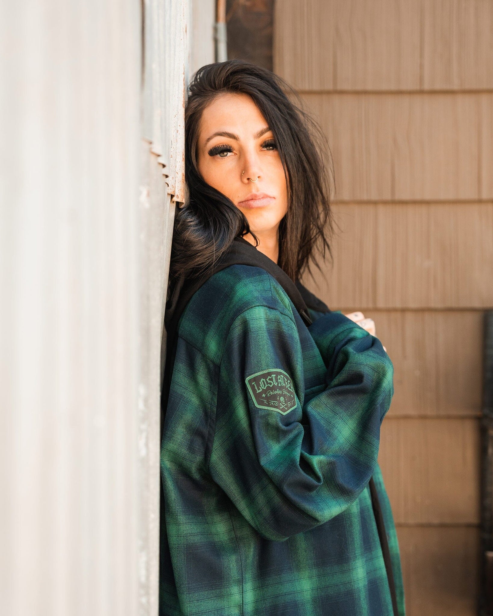 A woman with long dark hair leans against a wooden wall, wearing Relentless Betrayals Lost At Sea Premium Hooded Flannel in green and blue plaid. She gazes at the camera neutrally, effortlessly showcasing its unisex style.