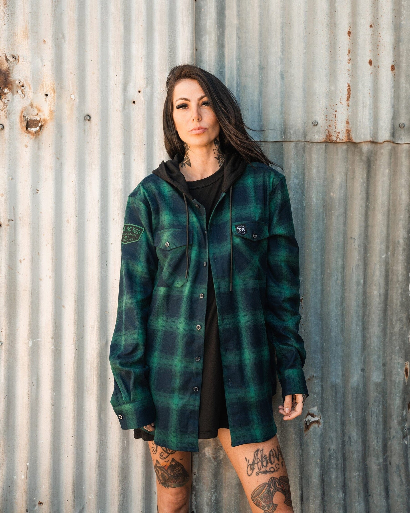 A tattooed person with long hair wears a green and black Lost At Sea Premium Hooded Flannel by Relentless Betrayal over a black hoodie, standing against a corrugated metal wall while looking at the camera.