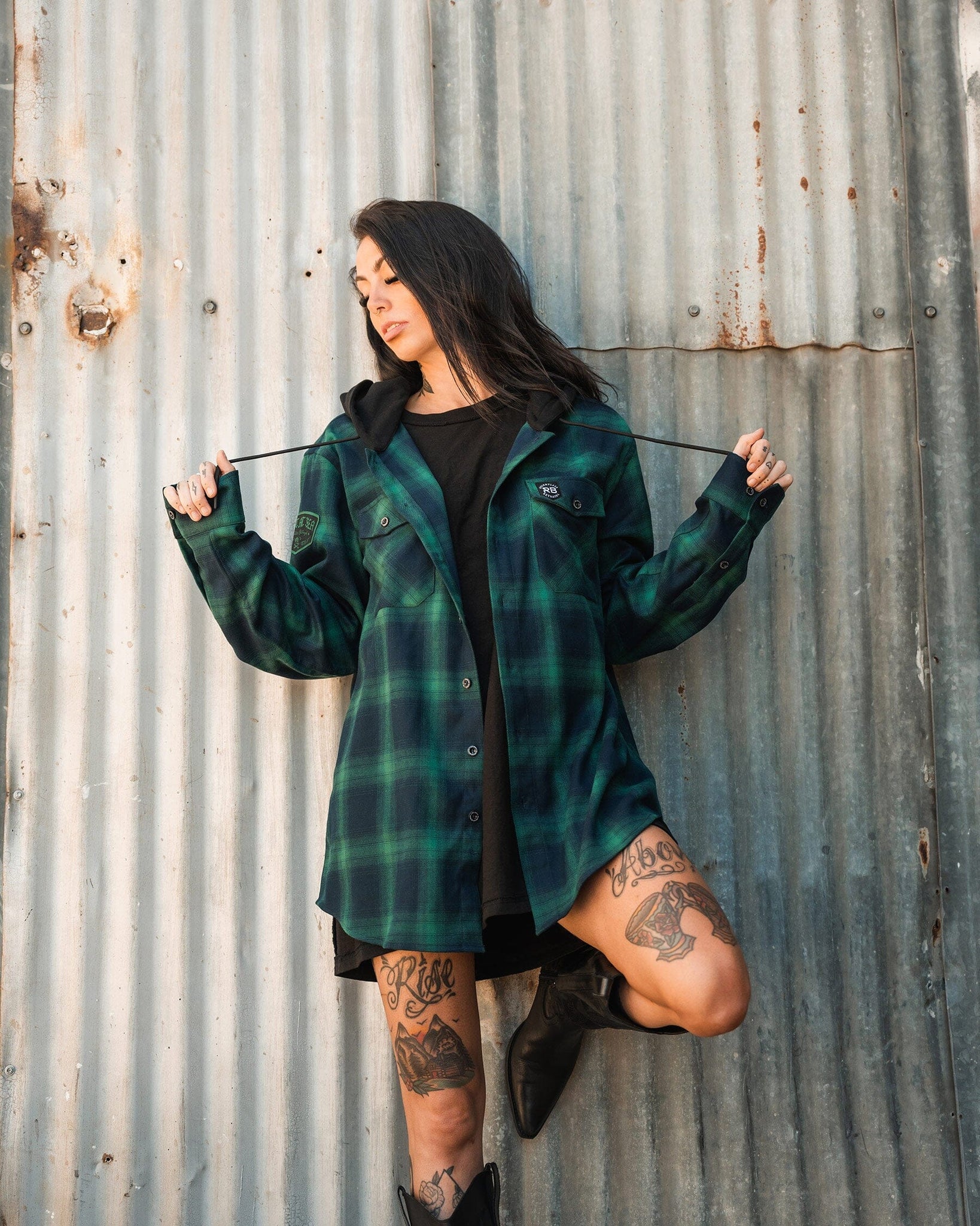 A person with long dark hair, wearing the Relentless Betrayal Lost At Sea Premium Hooded Flannel over a black outfit, stands against a corrugated metal wall. They have leg tattoos and are pulling on the hood strings while resting one foot on the wall in black boots.