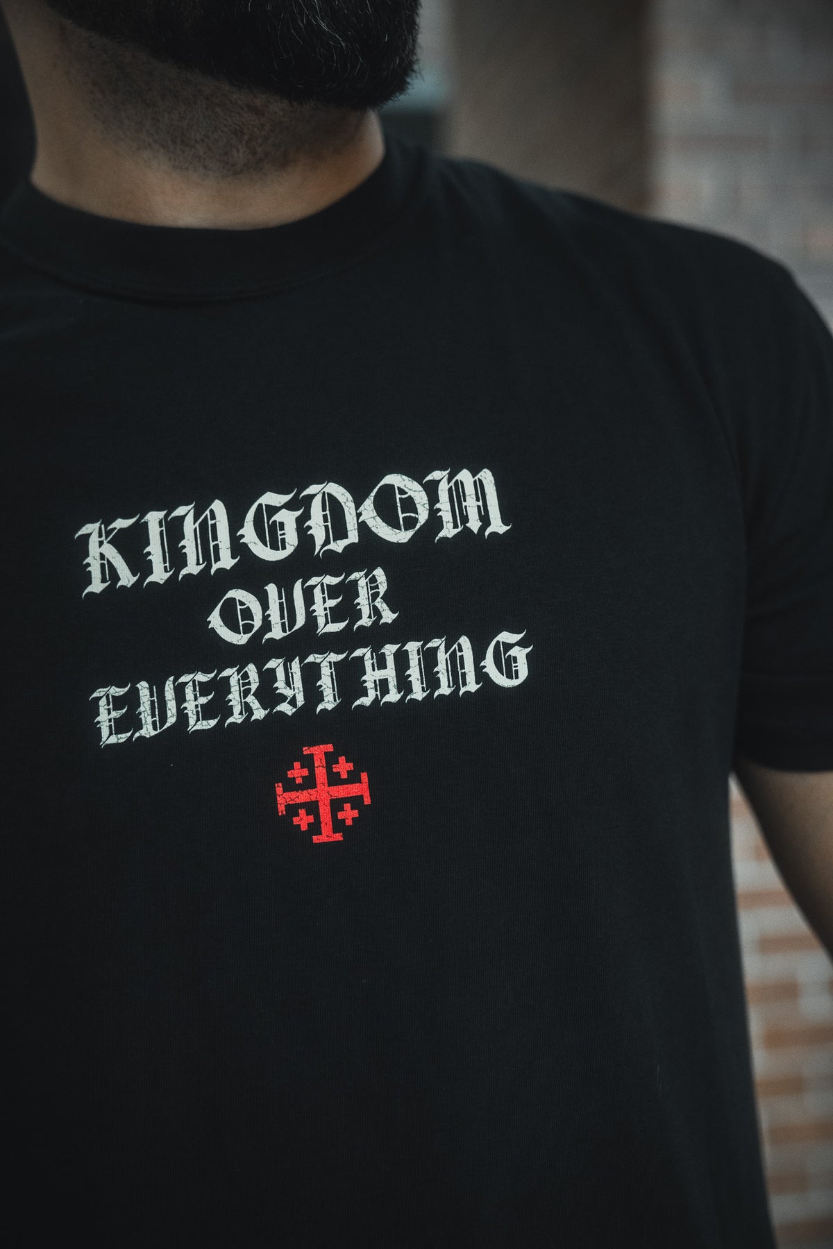 Someone wears the 13th Kingdom Over Everything Premium Tee—a black shirt with KINGDOM OVER EVERYTHING in bold white gothic font, a red cross below, inspired by Matthew 6:33 to remind us to seek first God’s Kingdom.