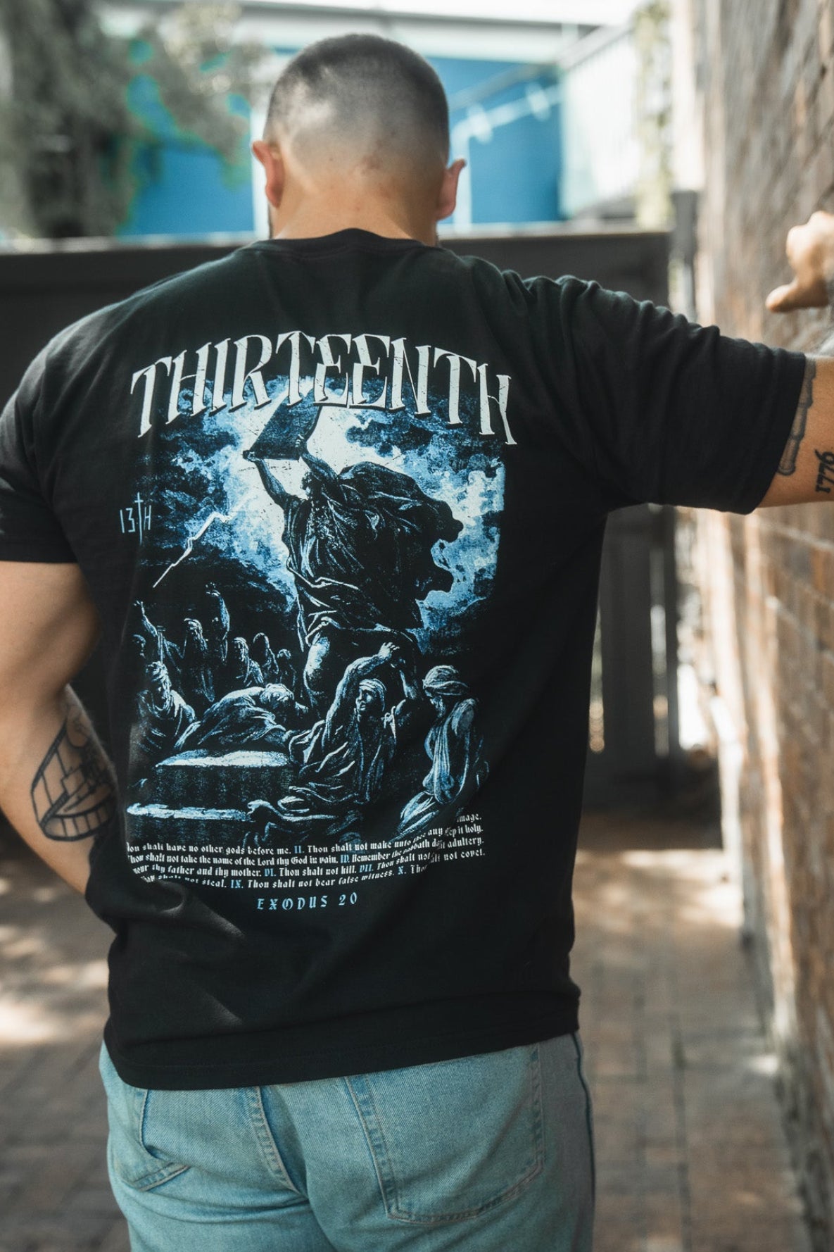 A person stands outdoors facing away, wearing the 13th brand’s The Ten Commandments Premium Tee, a black T-shirt with striking artwork, "THIRTEENTH" in bold above a biblical illustration and Ten Commandments text.