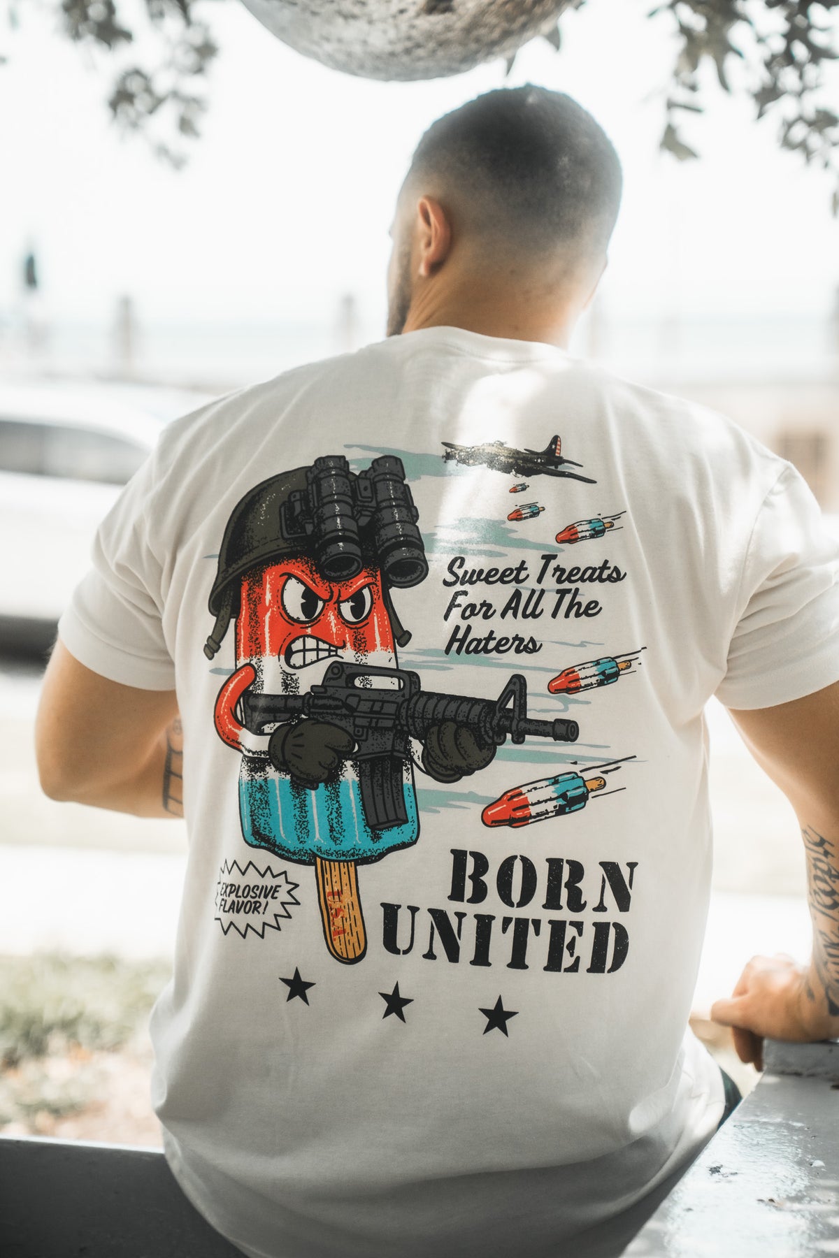 A man sits outdoors with his back to the camera, wearing a Born United Private Label "Sweet Victory" T-shirt featuring a cartoon popsicle with night vision goggles and fighter jets, with the text “Sweet Treats For All The Haters” and “BORN UNITED.”.