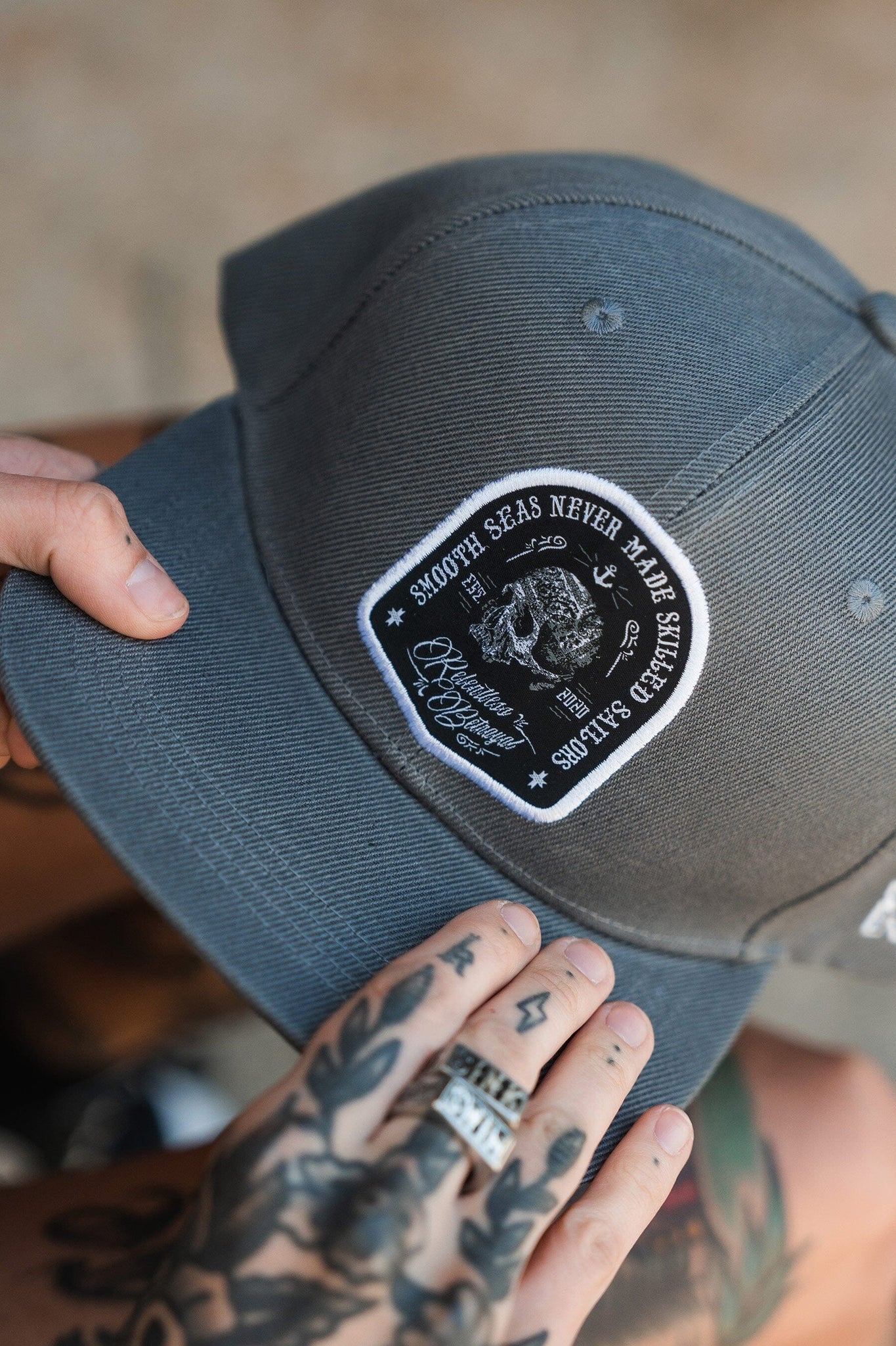 The Skilled Sailor Snapback by Relentless Betrayal is a gray hat made from a polyester-cotton blend. It features a black and white ship patch with Smooth seas never made a skilled sailor. Tattooed hands hold the cap, with a blurred shoe in the background.