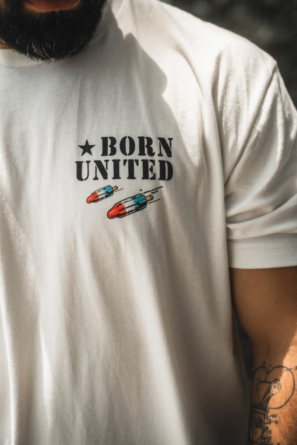 A person wearing the "Sweet Victory" white t-shirt by Born United Private Label, featuring BORN UNITED text and two small rockets on the chest. The image shows part of their beard and a tattooed arm.