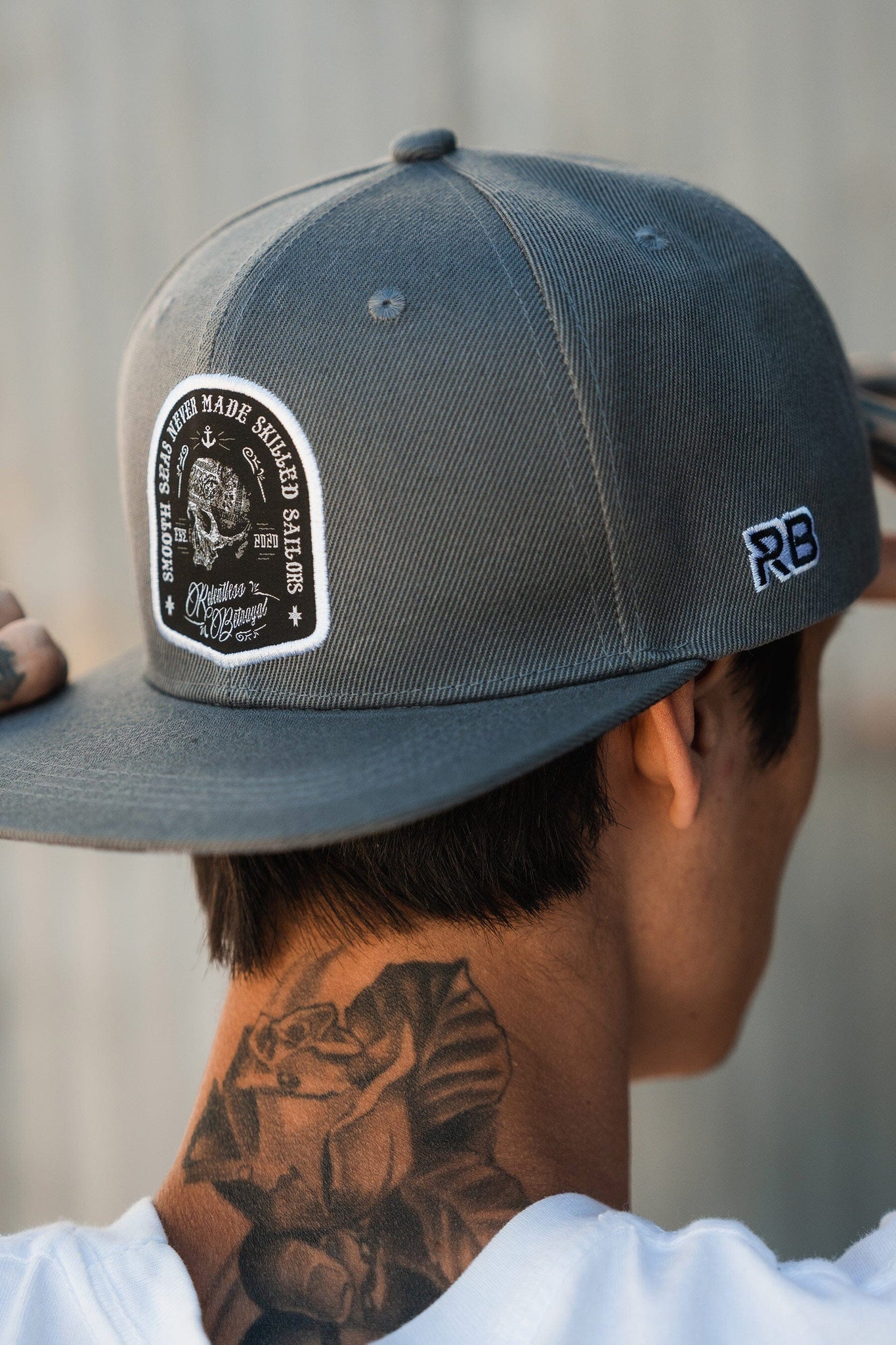 A person wearing the Skilled Sailor Snapback by Relentless Betrayal, featuring a motorcycle design and embroidered initials, is shown from the back. A rose tattoo adorns their neck as they adjust the stylish gray cap with detailed patch.