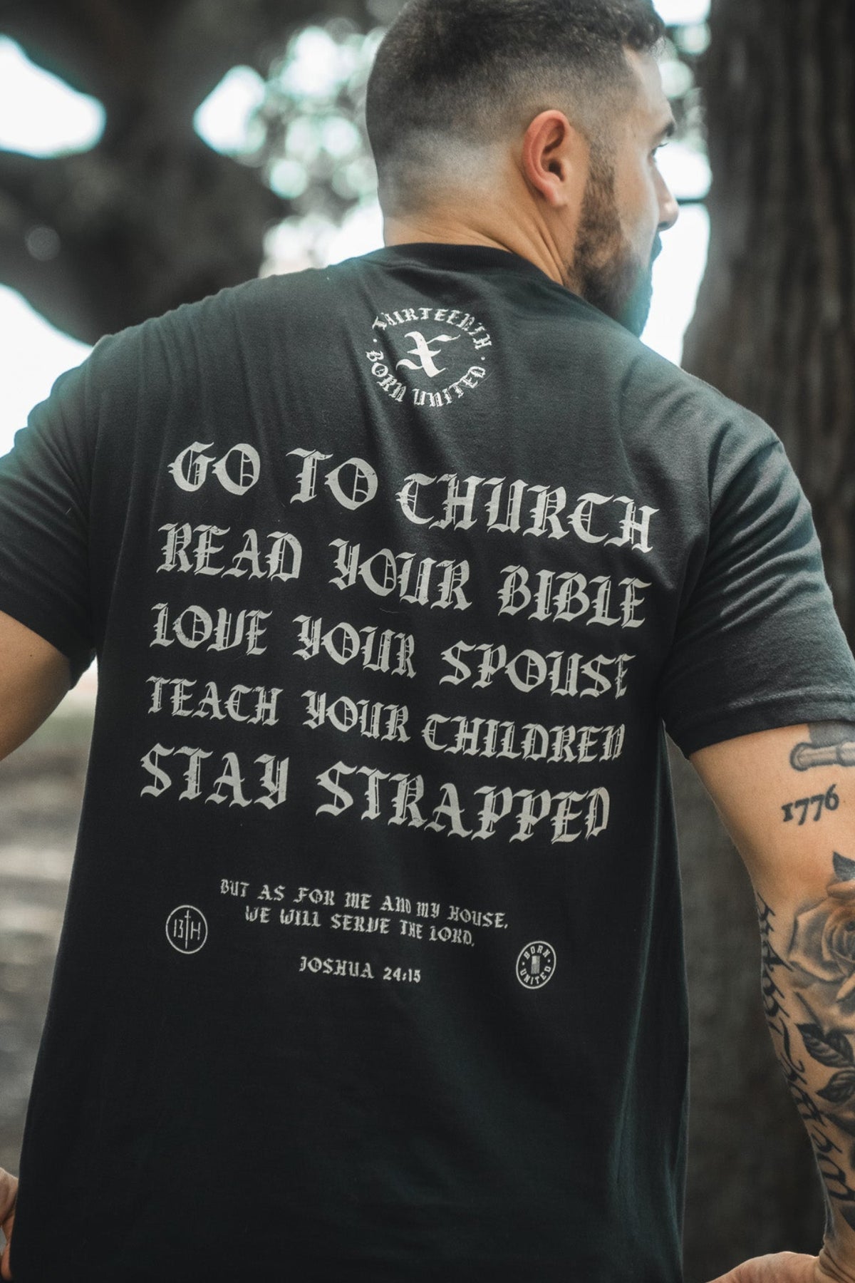 A bearded man outdoors wears the Kingdom Protector Premium Tee by Born United Private Label, featuring bold white text on the back: Go to church, Read your Bible, Stay strapped, and Joshua 24:15—ideal for Bible verse wall art inspiration.