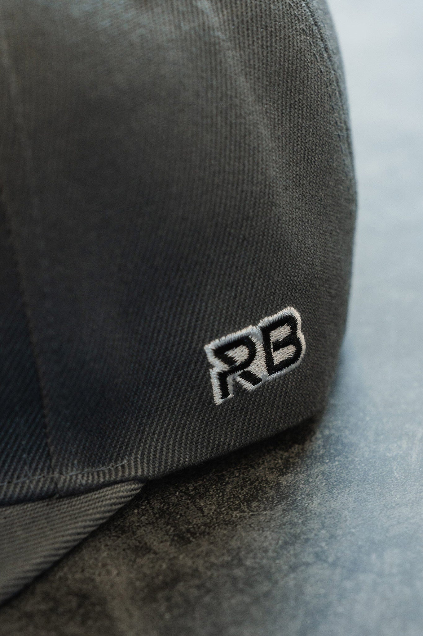 A close-up of the Skilled Sailor Snapback by Relentless Betrayal shows its dark gray fabric in detail. The polyester cotton blend highlights texture, and the embroidered white RB logo stands out on the side.