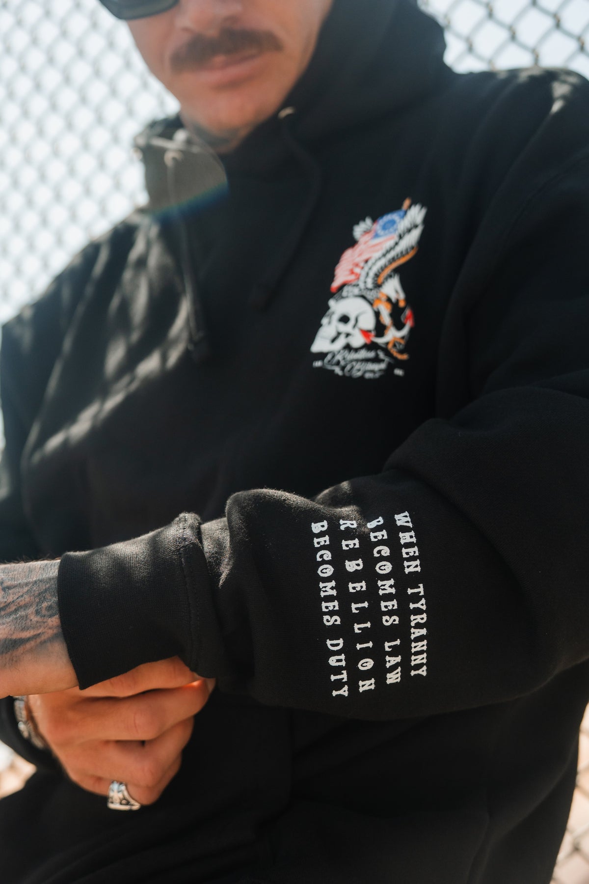 A person wears the Revolution Premium Hoodie by Relentless Betrayal, featuring a skull, eagle, and U.S. flag chest graphic with sleeve text: WHEN TYRANNY BECOMES LAW REBELLION BECOMES DUTY—styled with tattoos and sunglasses for bold integrity.