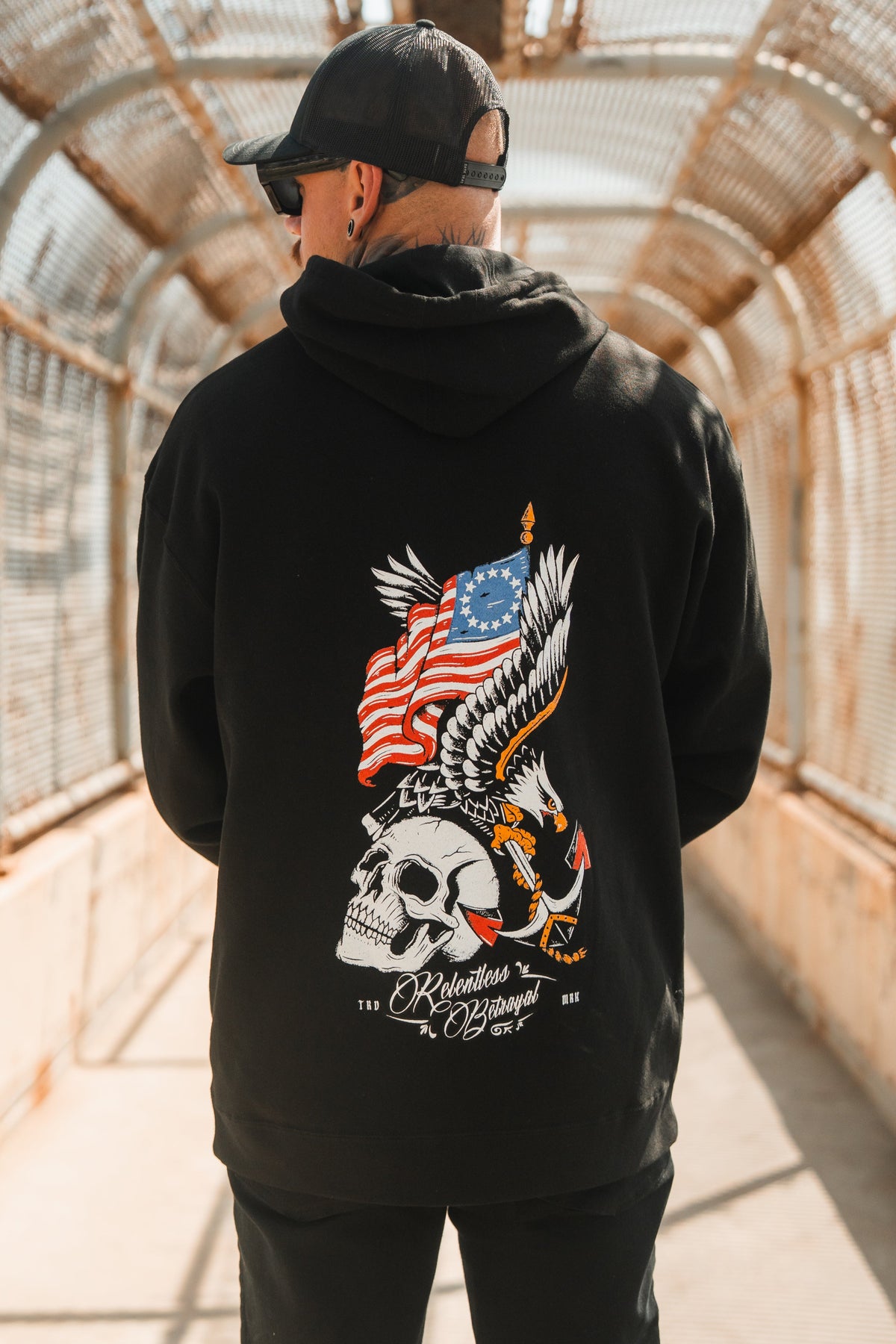 Wearing a black cap and the Revolution Premium Hoodie by Relentless Betrayal, a person stands on a sunlit bridge. The hoodie’s back displays a skull, eagle, American flag, roses, and “Relentless Royal”—a bold statement of integrity.