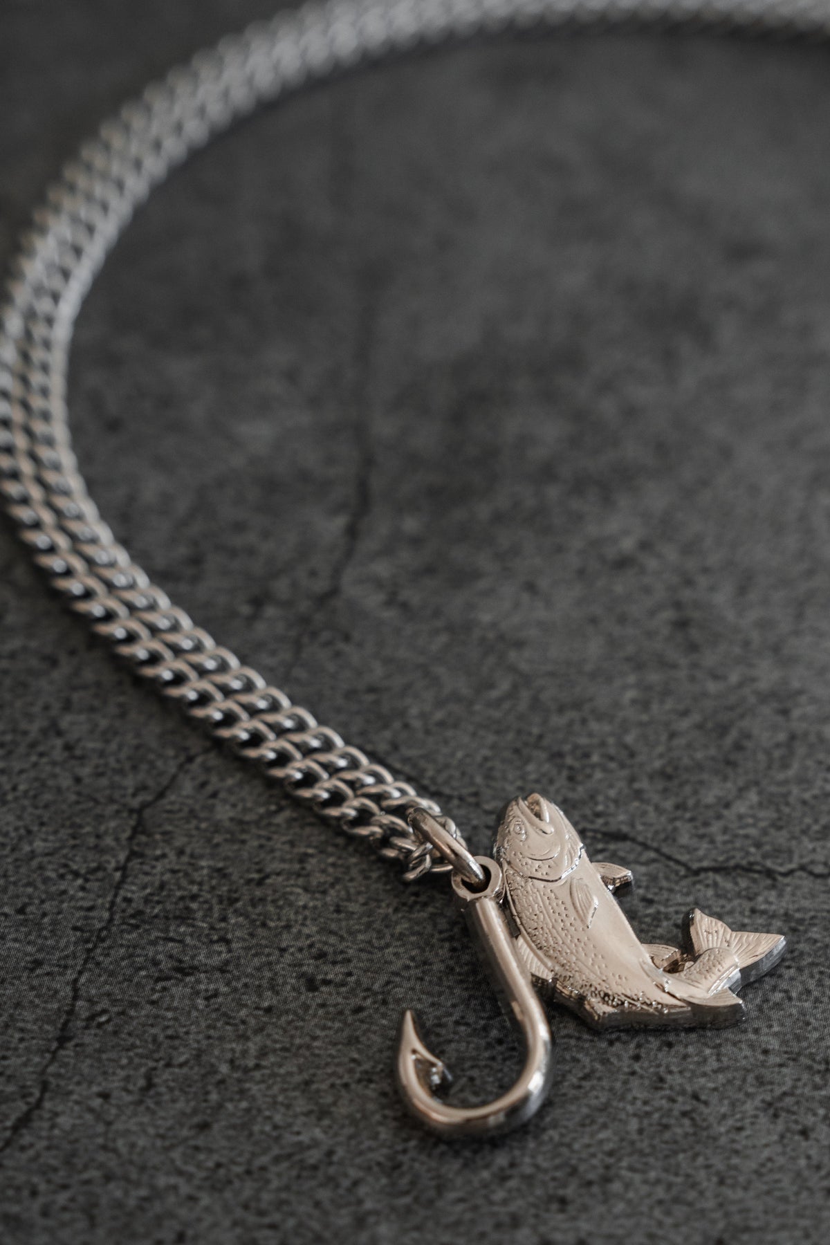 The Mucker Bucket Fly Fishin’ Trout Hunter – Stainless Steel Necklace, featuring a silver trout and hook pendant, rests on a textured gray surface, suspended from a durable stainless steel chain.