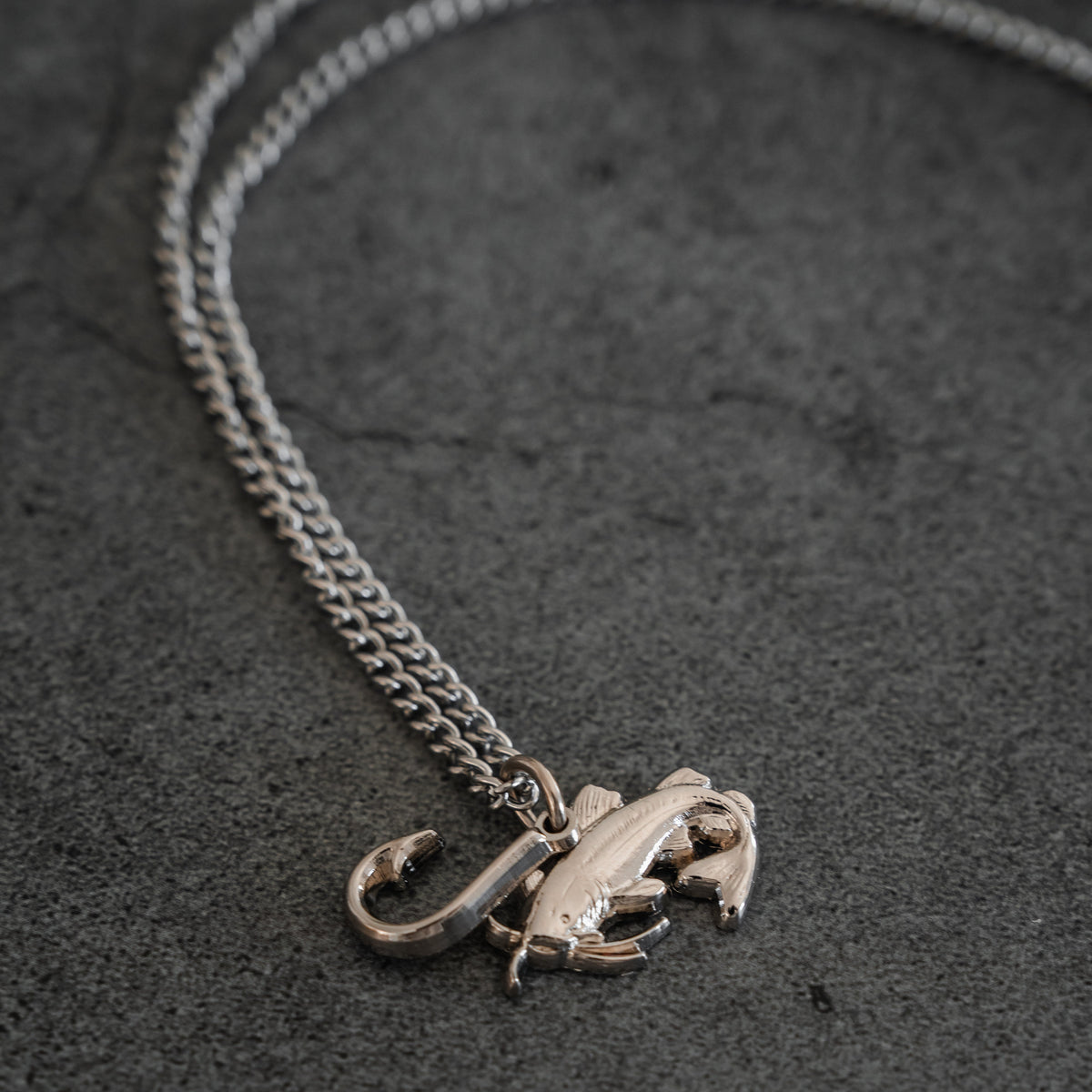 The Mud Cat Noodler Stainless Steel Necklace by Mucker Bucket features a silver chain with a fish and fish hook pendant, crafted in the USA and shown on a dark grey stone surface.