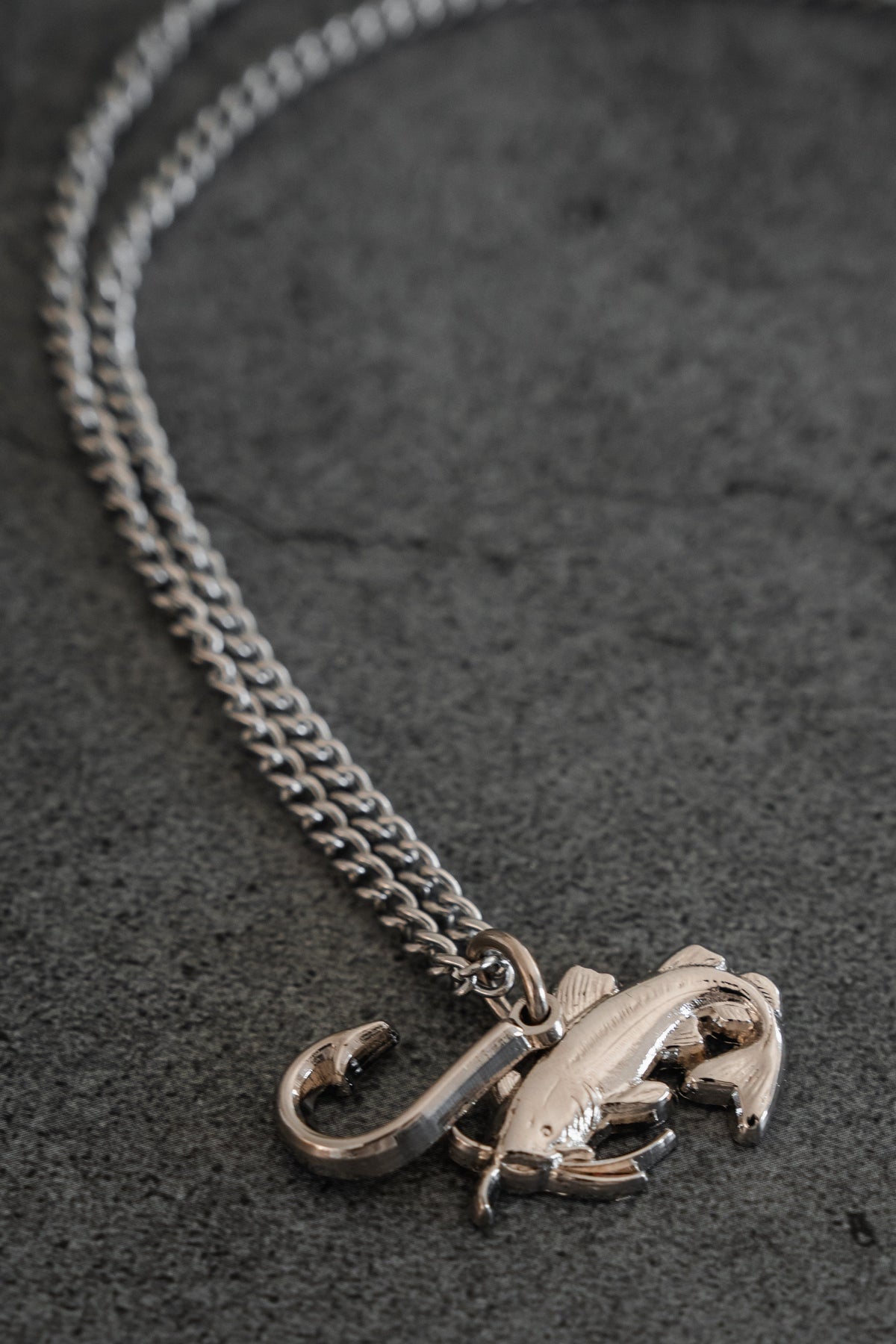 The Mud Cat Noodler Stainless Steel Necklace by Mucker Bucket features a silver chain with a fish and fish hook pendant, crafted in the USA and shown on a dark grey stone surface.