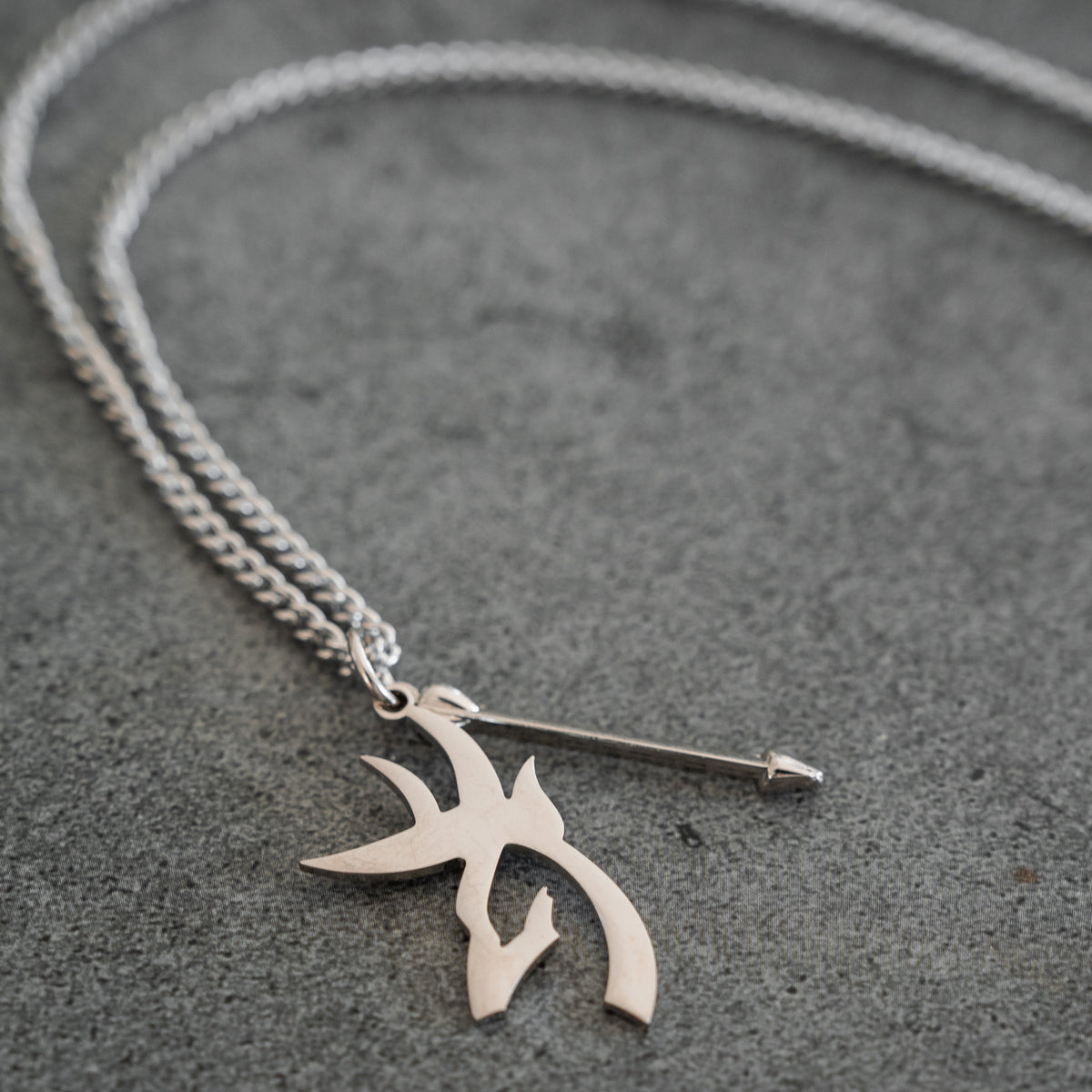The Mucker Bucket Big Buck Bow Hunter - Stainless Steel Necklace, featuring a chain link with two pendant charms—one shaped like an arrow and the other adorned with an abstract, tribal-inspired design—rests on a gray surface. This distinctive piece includes a stainless steel pendant that celebrates the spirit of the Bow Hunter and is proudly made in the USA.