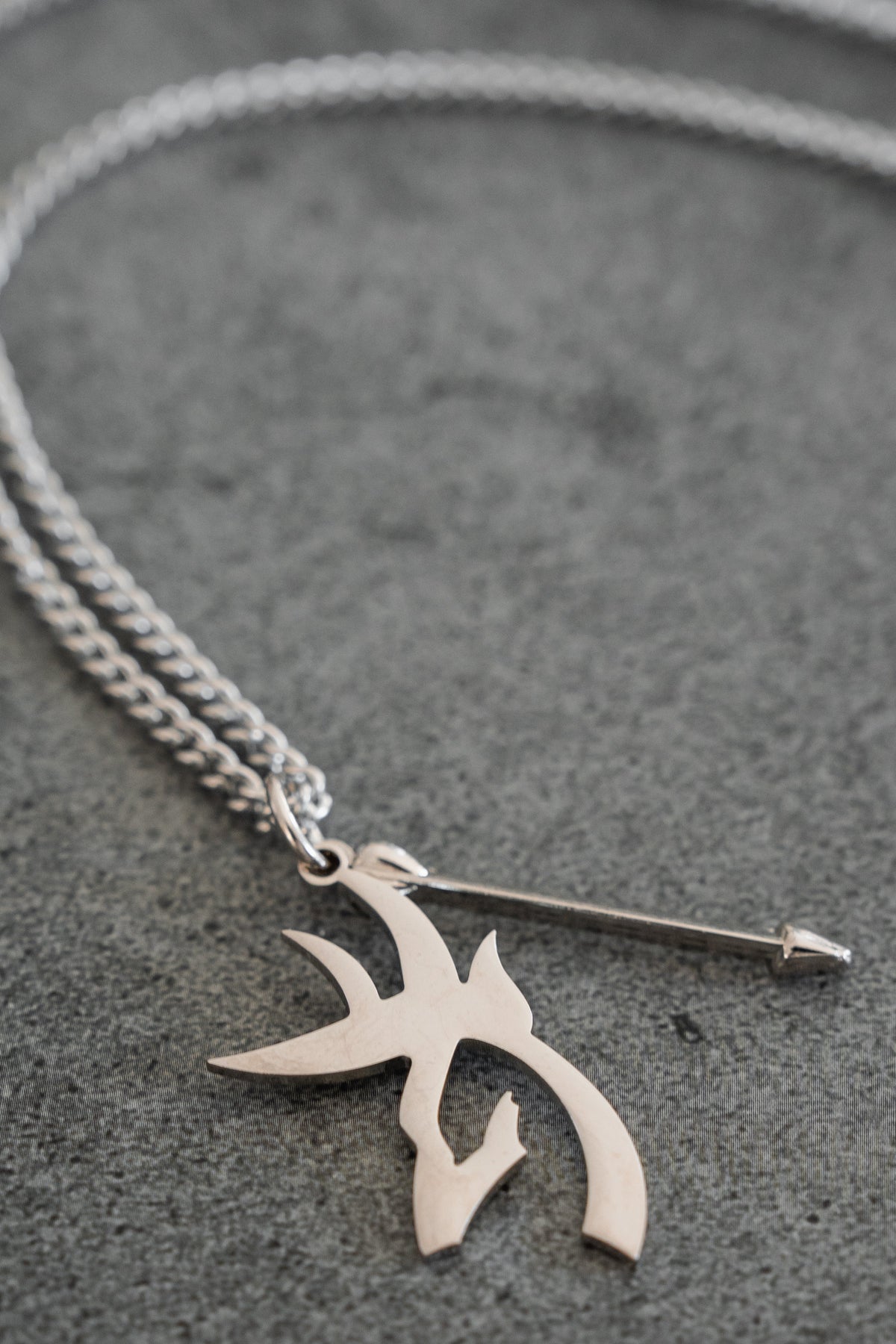 The Mucker Bucket Big Buck Bow Hunter - Stainless Steel Necklace, featuring a chain link with two pendant charms—one shaped like an arrow and the other adorned with an abstract, tribal-inspired design—rests on a gray surface. This distinctive piece includes a stainless steel pendant that celebrates the spirit of the Bow Hunter and is proudly made in the USA.