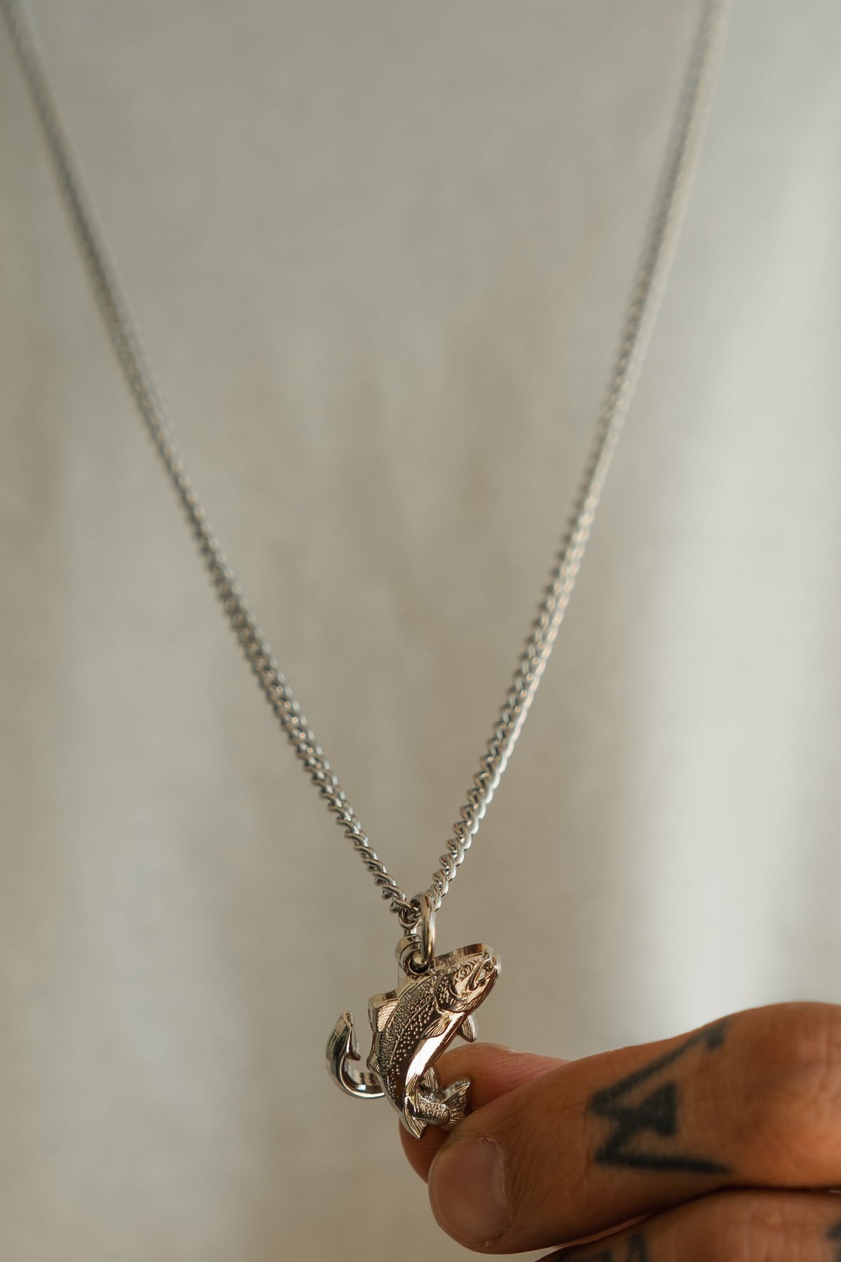 A tattooed hand holds the Mucker Bucket Fly Fishin’ Trout Hunter Stainless Steel Necklace, featuring a detailed trout pendant on a stainless steel chain, with a white shirt visible in the background.