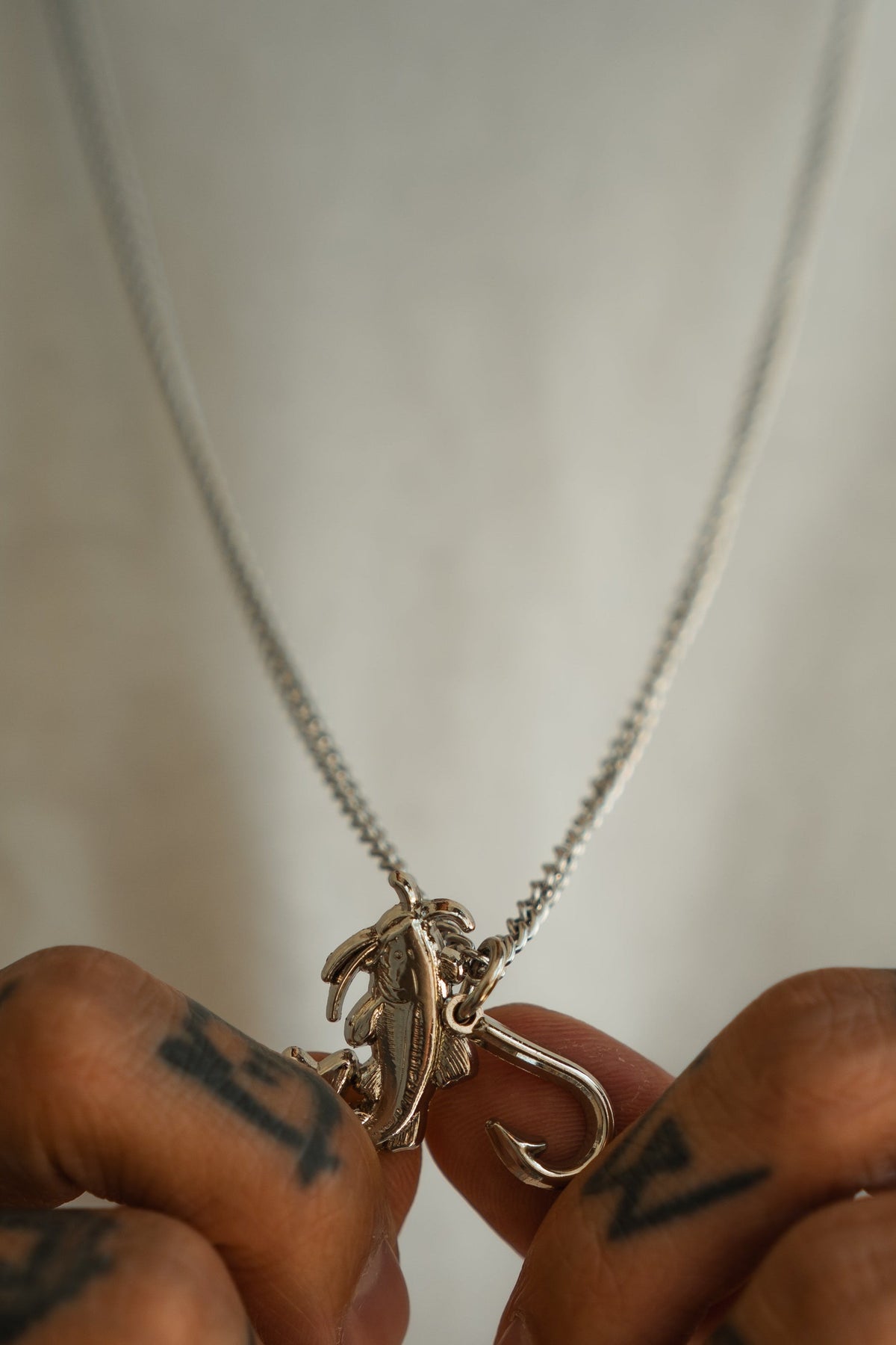 A person with tattooed fingers holds the Mud Cat Noodler - Stainless Steel Necklace by Mucker Bucket, which features a stainless steel fish and hook pendant suspended from a matching chain, displayed against a plain white shirt background.