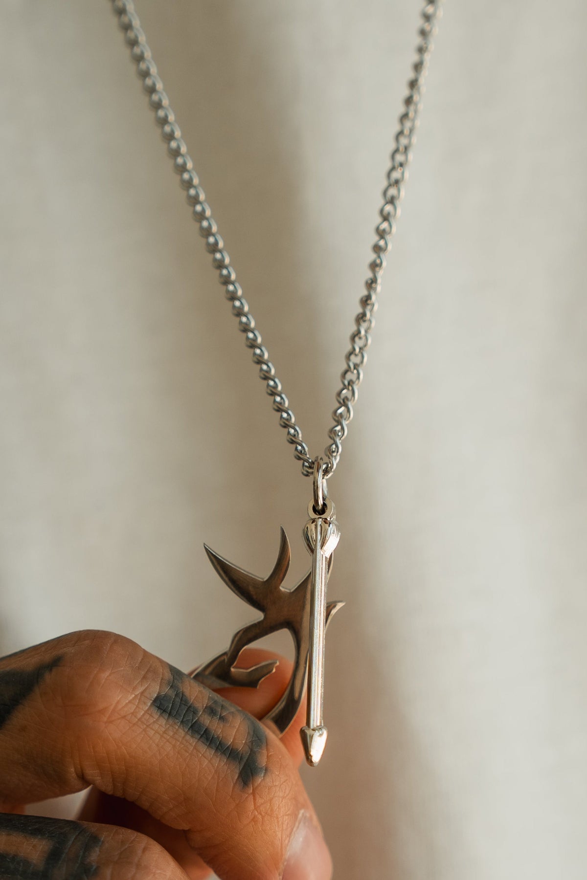 A hand with tattoos holds the Mucker Bucket Big Buck Bow Hunter Stainless Steel Necklace, featuring a silver pendant with a stylized arrow and abstract design inspired by the spirit of the bow hunter, set against a plain light background.