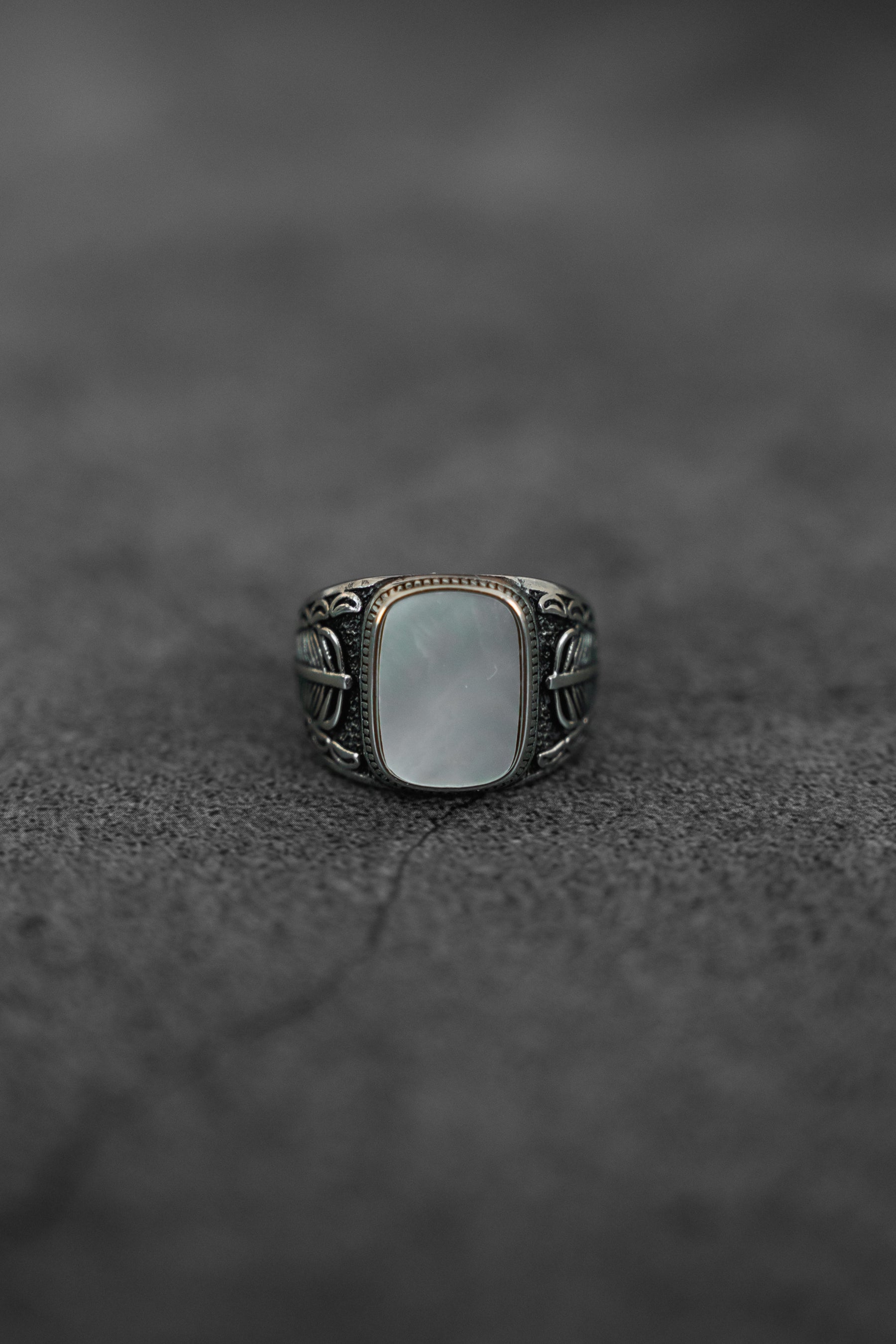 The Desire Opal Ring by Relentless Betrayal, featuring a large rectangular white opal and an ornate handcrafted band, is centered on a textured dark surface.