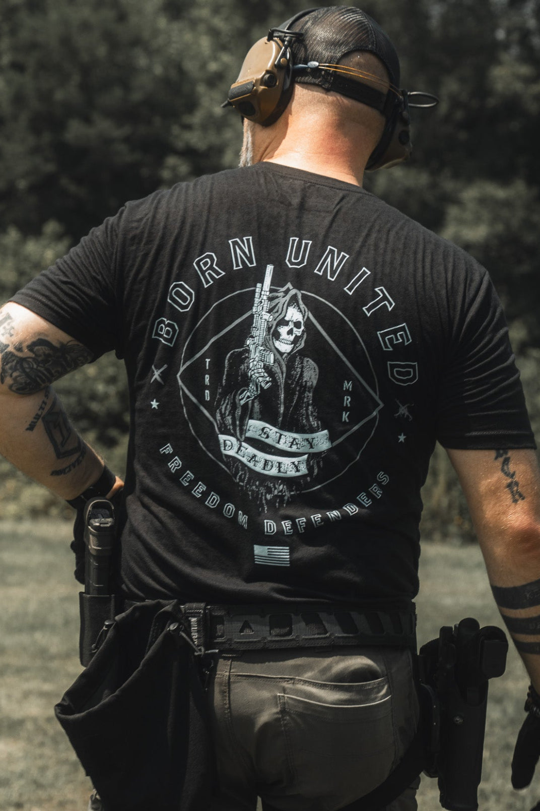 A man wearing tactical gear and ear protection stands outdoors, displaying the back of his black Stay Deadly Tee by Born United Private Label—a t-shirt featuring a graphic of a woman holding a gun with the text "Destroy Deceit," capturing the bold spirit of patriots.
