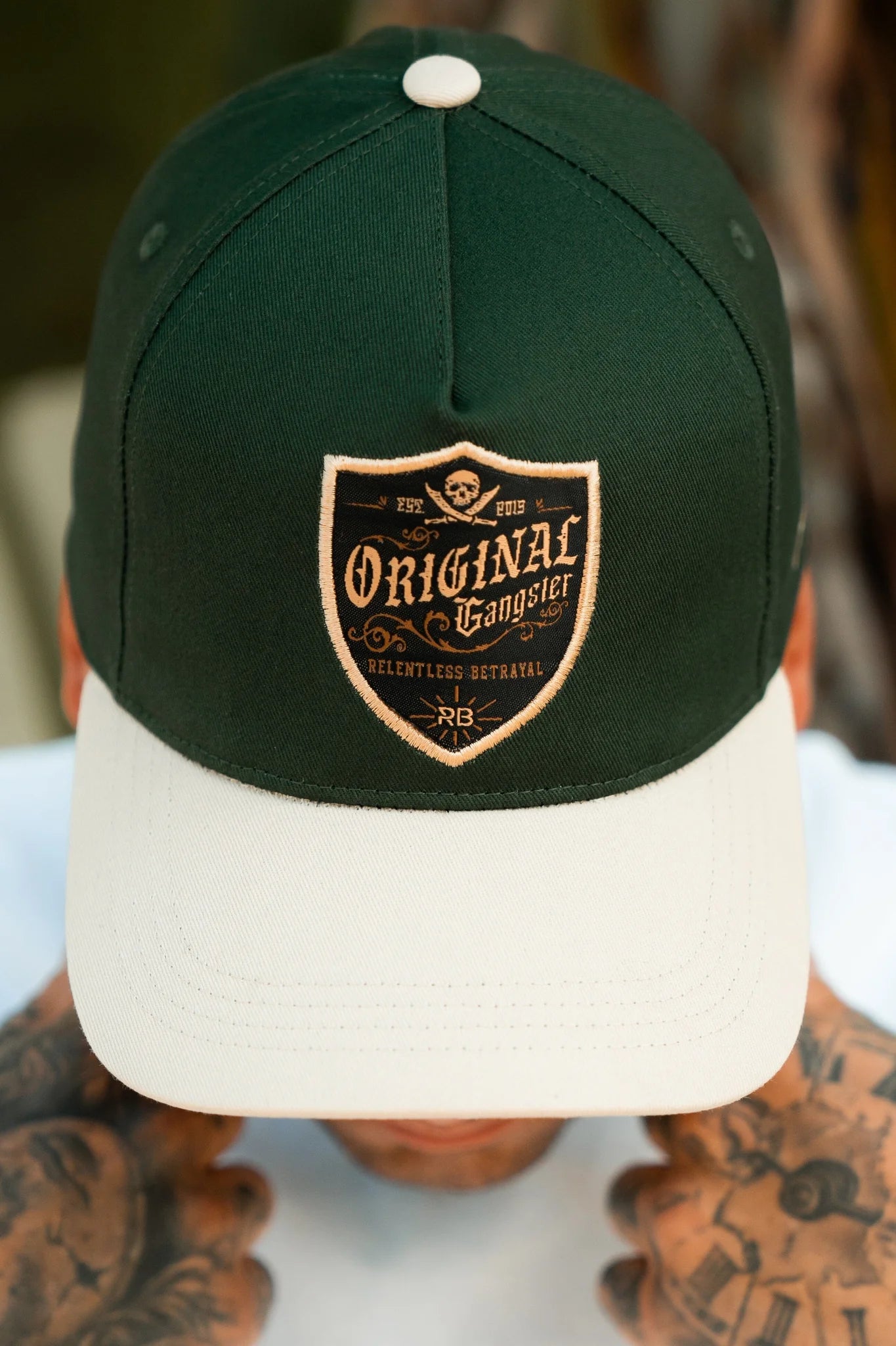 A person is wearing a Relentless Betrayal OG Forest Green Snapback, featuring a beige brim and Original Gangster patch. Their tattooed hands rest on their cheeks against a blurred background.