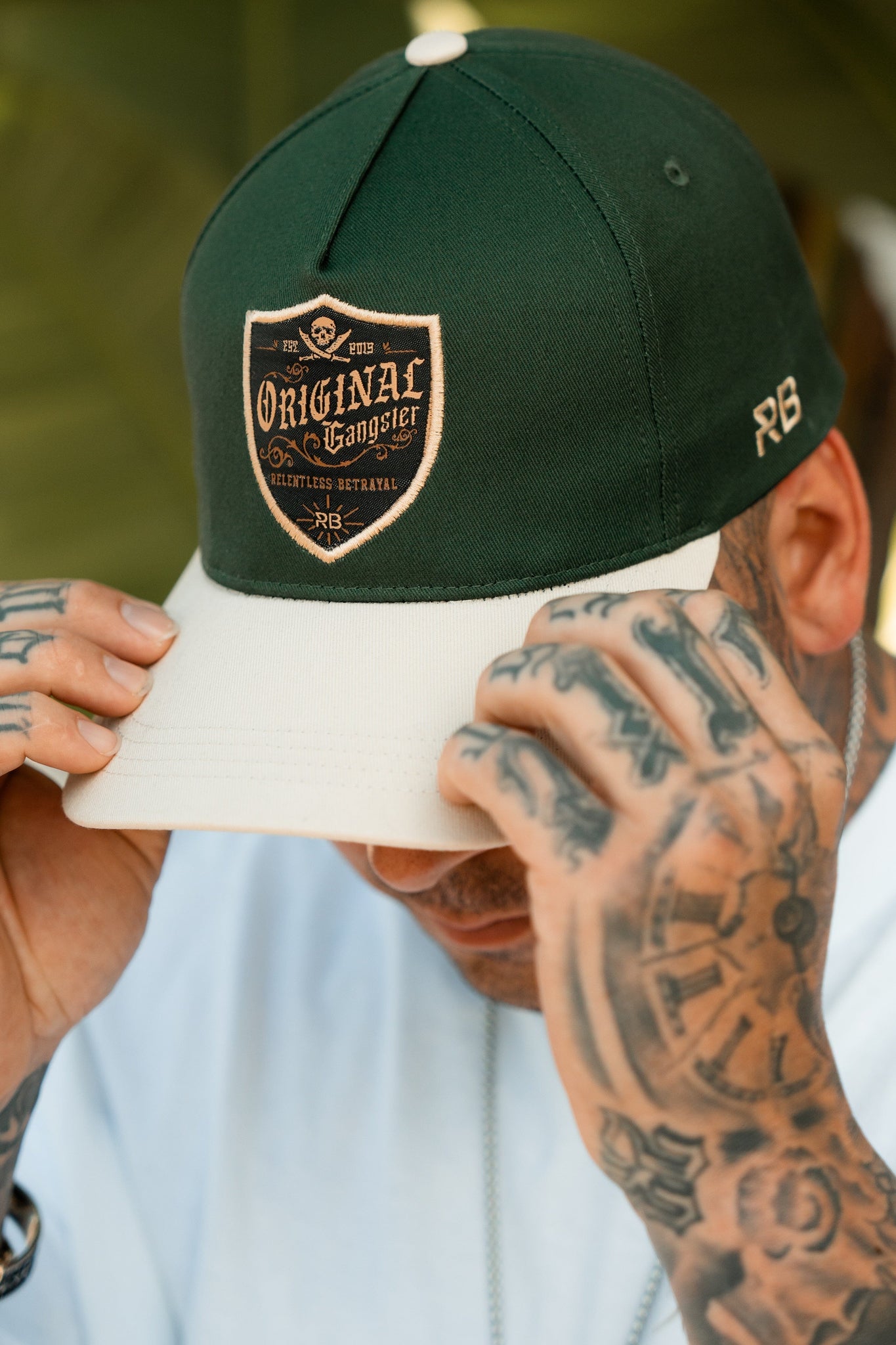 A person with tattooed hands adjusts the brim of a Relentless Betrayal OG Forest Green Snapback. The cap, featuring a patch that reads Original Gangster amid small designs, complements their light-colored shirt against a blurred background.
