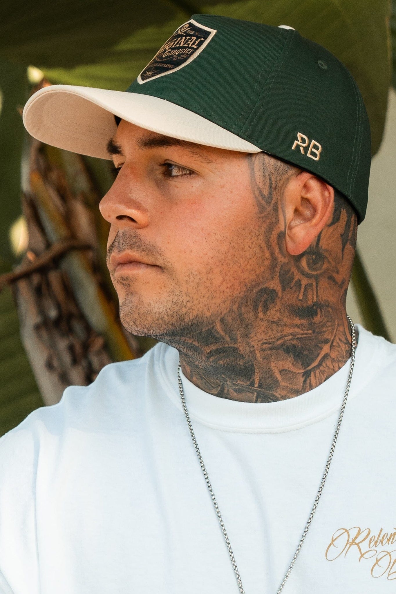 A man with tattoos on his neck and face gazes to the side, wearing a Relentless Betrayal OG Forest Green Snapback with an embroidered design and curved brim, paired with a white T-shirt. A silver chain adorns his neck against the backdrop of blurred foliage.