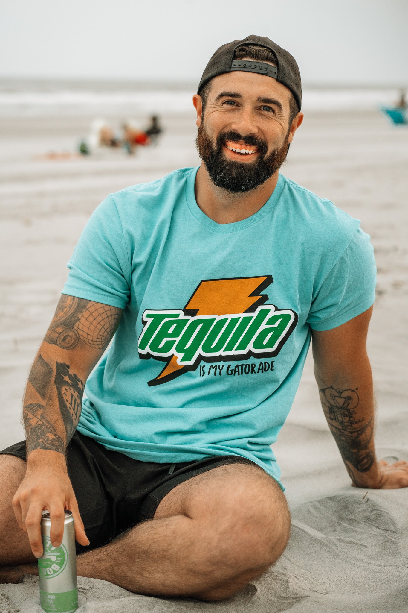 A bearded, tattooed man smiles on the beach with a can in hand, wearing Tango Charlie Apparel’s “Tequila Is My Gatorade - Berry Blue” shirt. People and the ocean blur behind him, capturing the perfect summer vibe.