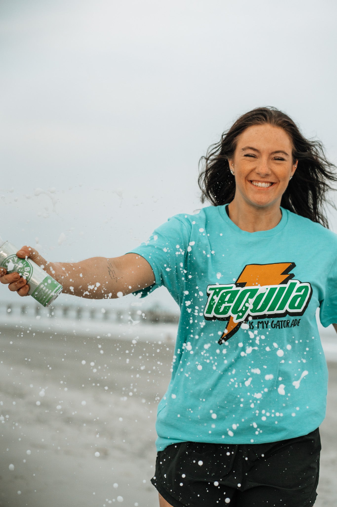 A smiling woman in a Tequila Is My Gatorade - Berry Blue shirt by Tango Charlie Apparel splashes water from a bottle on a summer beach, with bold colors and a blurred ocean and pier in the background.