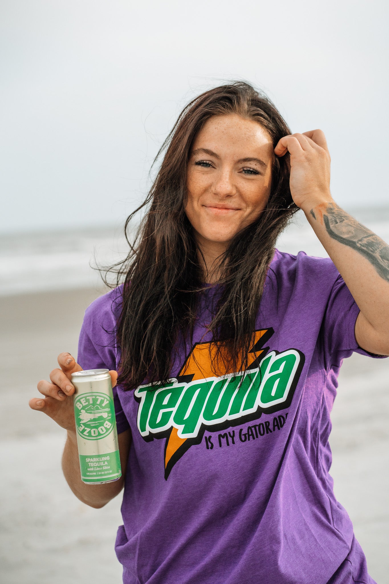 A woman with long dark hair stands on a beach, smiling in a Tango Charlie Apparel “Tequila Is My Gatorade - Purple Rush” t-shirt as she holds a green can, with the ocean and sky behind her.