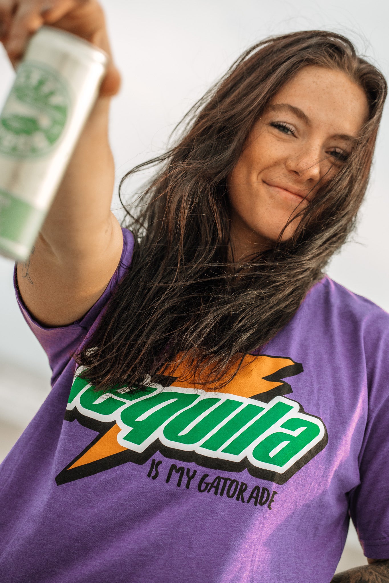 A woman with long brown hair smiles and holds a can out to the camera, wearing Tango Charlie Apparel’s bold purple “Tequila Is My Gatorade - Purple Rush” shirt.