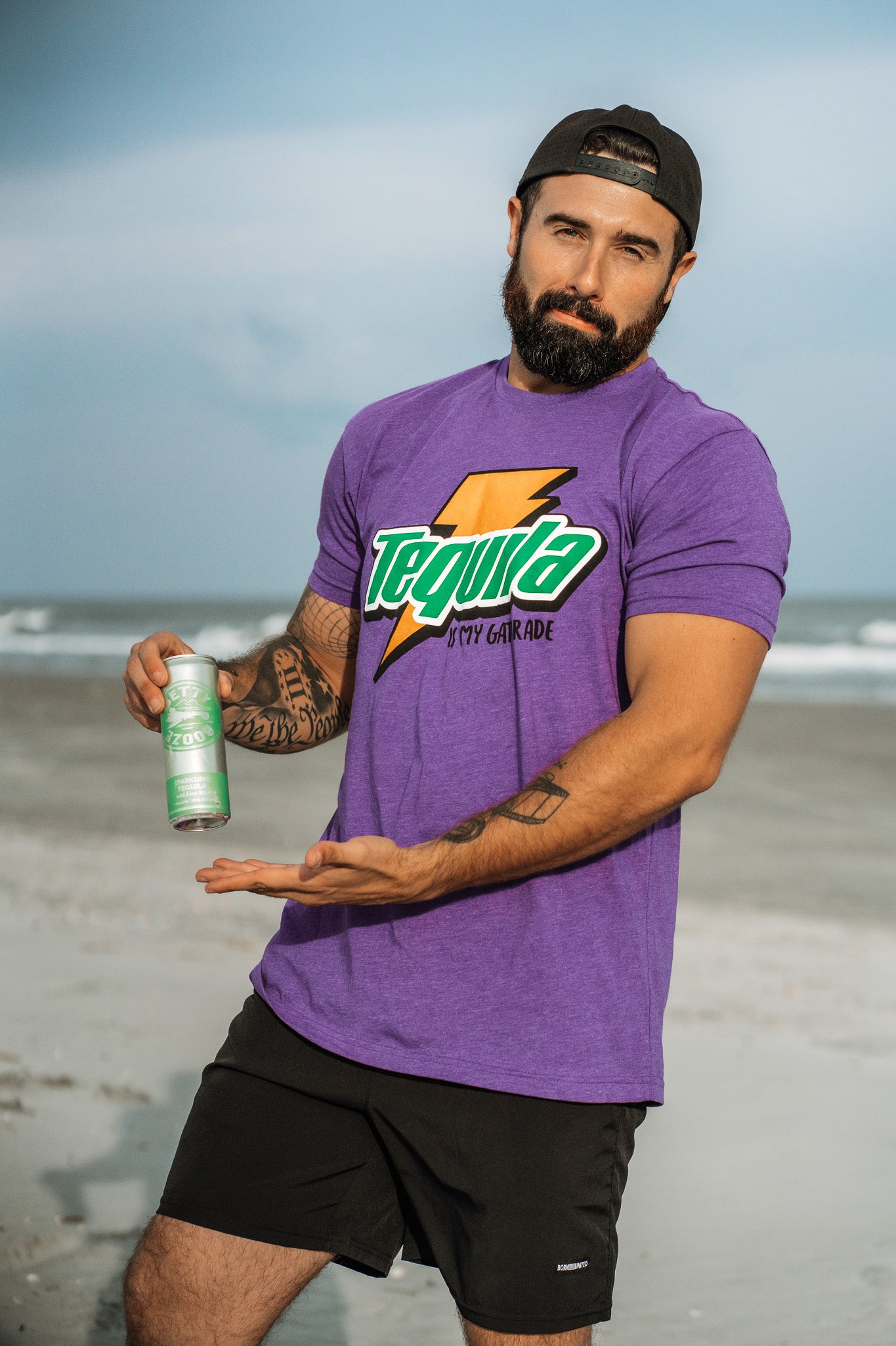 A bearded man at the beach sports a bold Tequila Is My Gatorade - Purple Rush T-shirt by Tango Charlie Apparel and black shorts, holding up a green can. He wears his black cap backwards and gestures toward the drink.