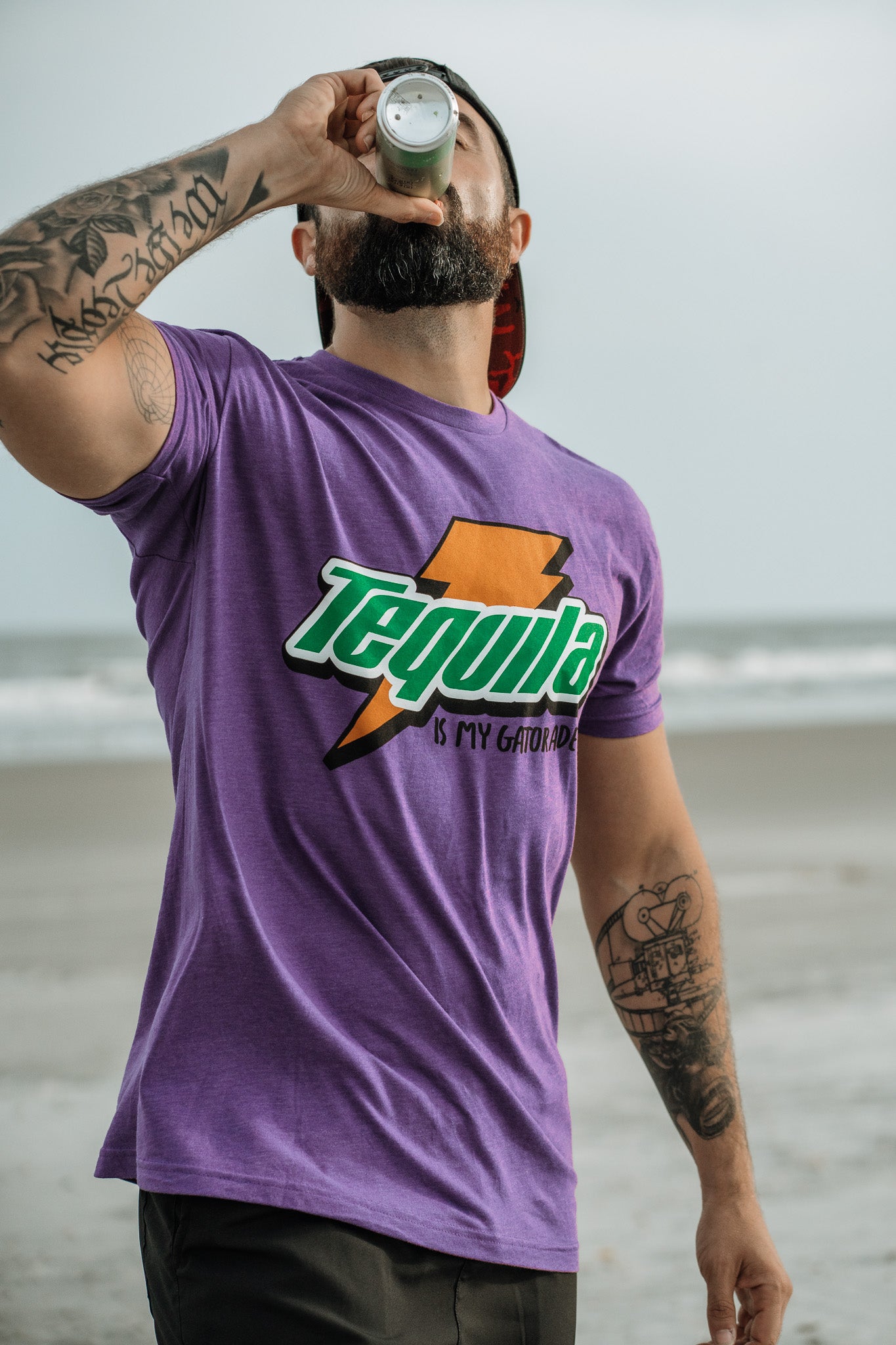 A tattooed man drinks from a bottle on the beach, wearing Tango Charlie Apparel’s “Tequila Is My Gatorade - Purple Rush” T-shirt as the ocean and sky provide a vivid backdrop.