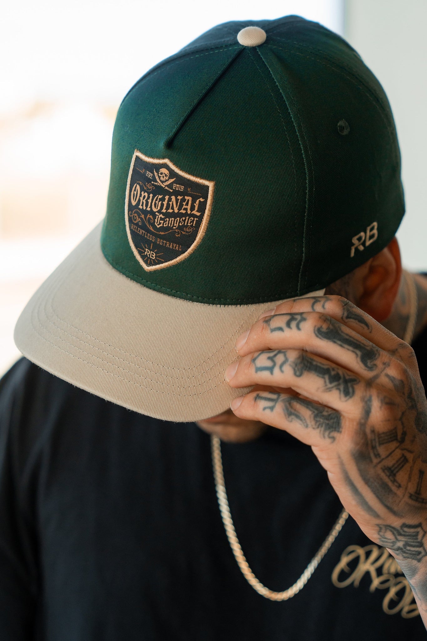 Wearing a Relentless Betrayal OG Forest Green Snapback featuring an Original Gangster patch, a person with tattooed hands pairs it with a black shirt and silver chain. The indoor lighting subtly enhances their style.