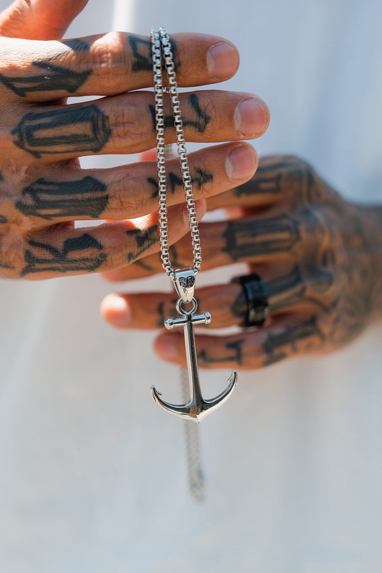 A tattooed hand grips the handcrafted Anchor Drop Necklace by Relentless Betrayal, featuring a large stainless steel anchor pendant on a 26-inch chain, set against a blurred background.