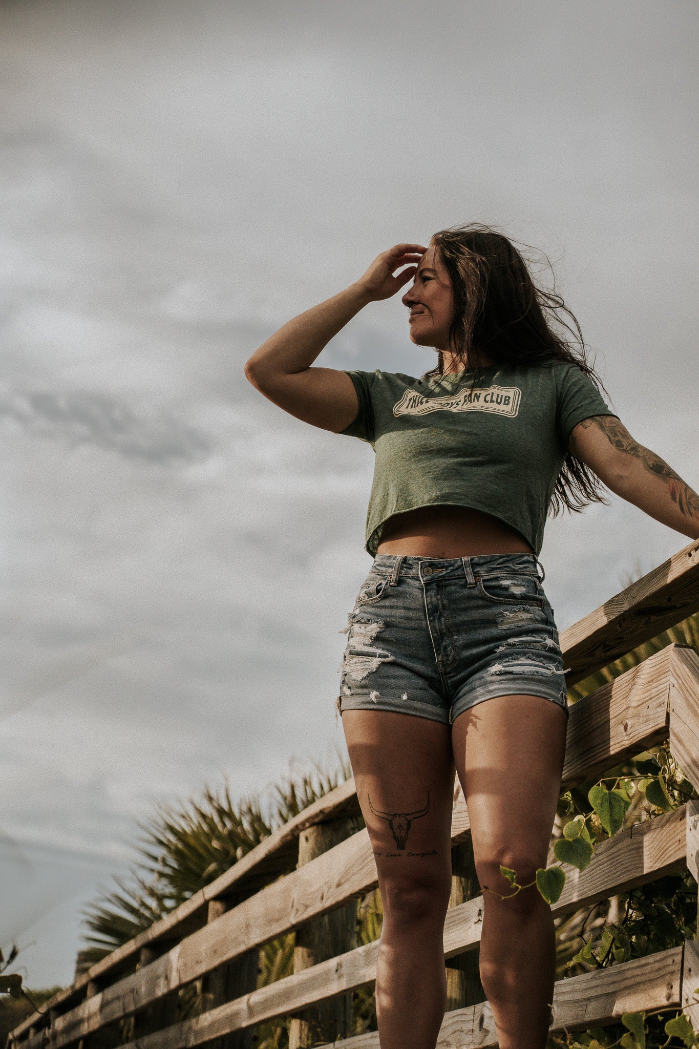 A woman in the THICC Boys Fan Club Crop T-Shirt by Tango Charlie Apparel and denim shorts stands on a wooden walkway outdoors, smiling and shielding her eyes as she looks into the distance beneath a cloudy sky.
