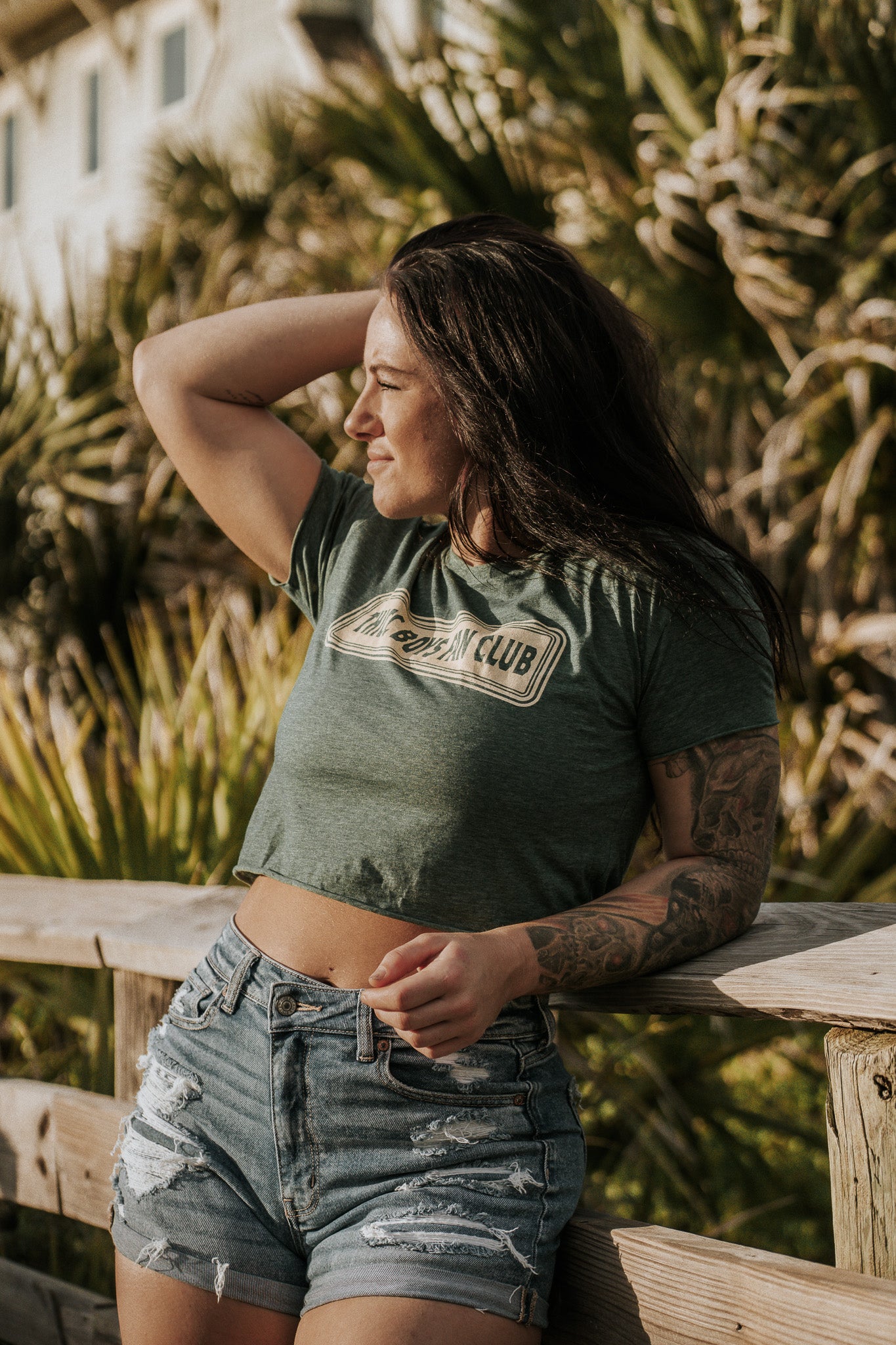 A woman with long dark hair stands outdoors on a wooden railing, wearing a green THICC Boys Fan Club Crop T-Shirt from Tango Charlie Apparel and ripped denim shorts, gazing into the distance under sunlight with greenery behind her.