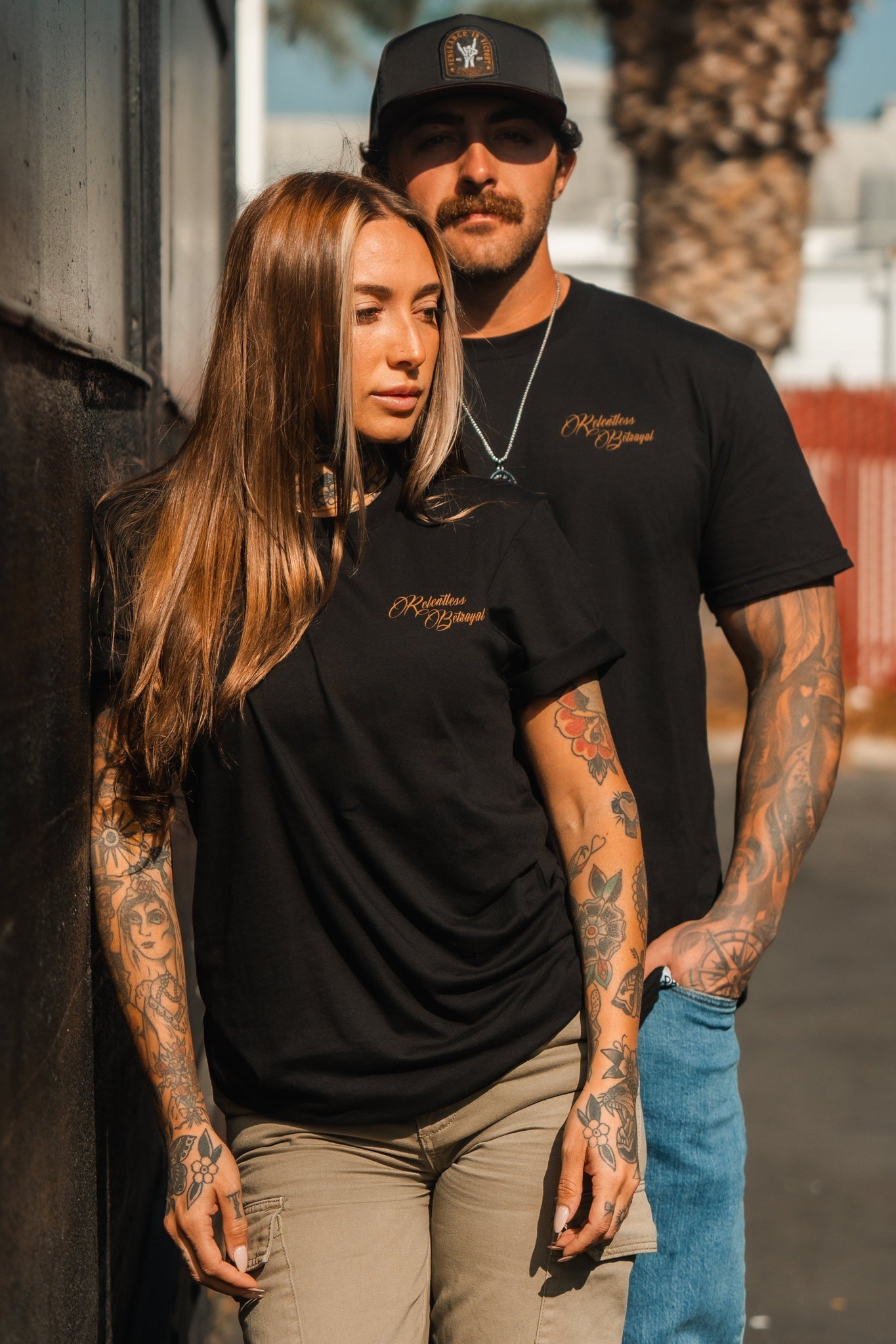A tattooed woman with long brown hair leans against a wall wearing a Vengeance Tee from Relentless Betrayal and beige pants, embodying resilience. Beside her, a tattooed man with a mustache and cap wears the same shirt and blue jeans. Theyre outside with a blurred background.