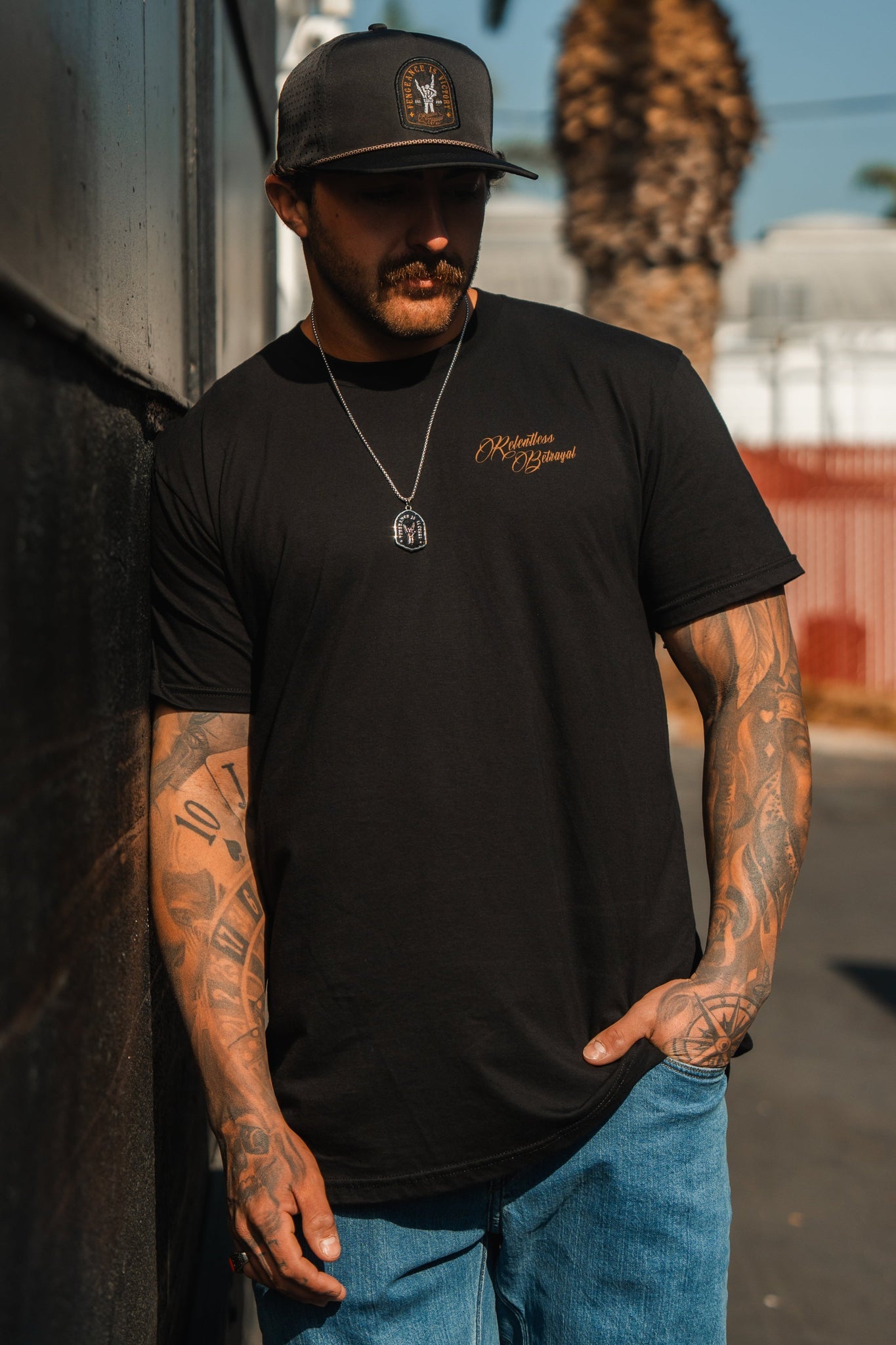 A man in a Vengeance Tee by Relentless Betrayal and jeans leans against an outdoor wall, emanating resilience and determination. He wears a cap, necklace, and has tattoos on his arms as a blurred palm tree gently sways in the background.
