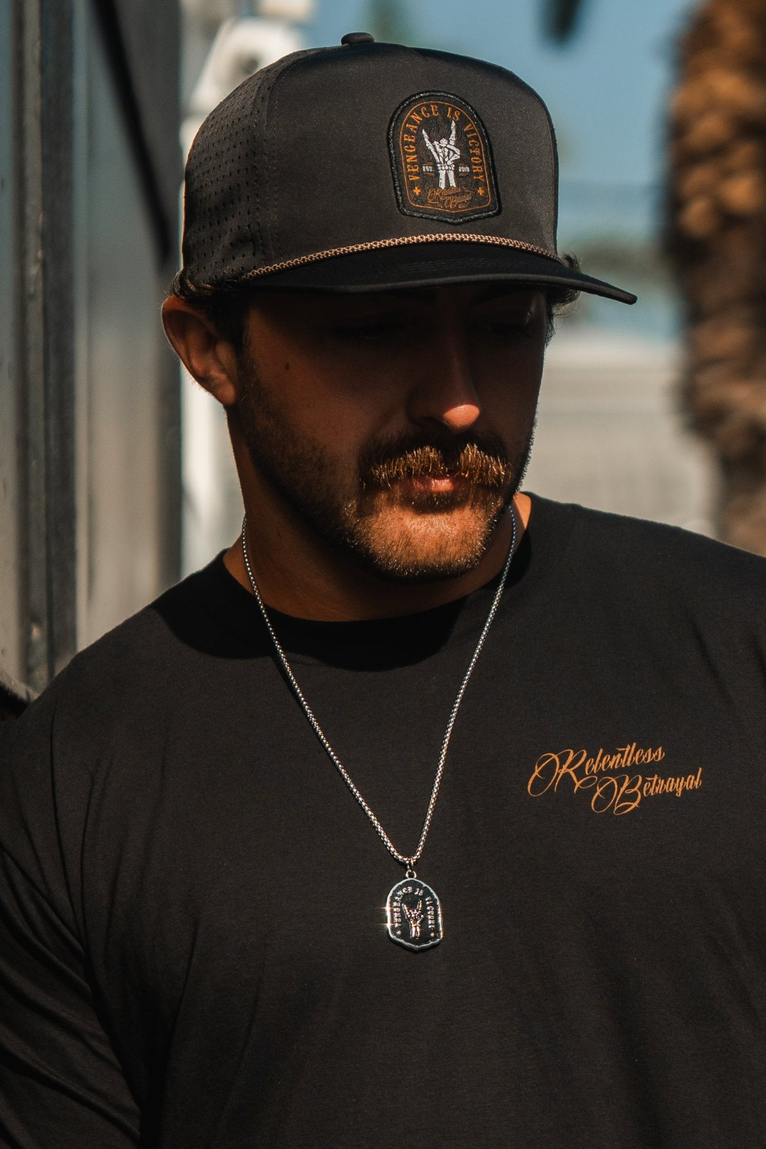 A man in a black cap with a patch and a shirt by Relentless Betrayal, inscribed Relentless Betrayal for resilience, stands outside. He wears the Vengeance Necklace featuring a rectangular pendant. The background is slightly blurred, hinting at a palm tree.