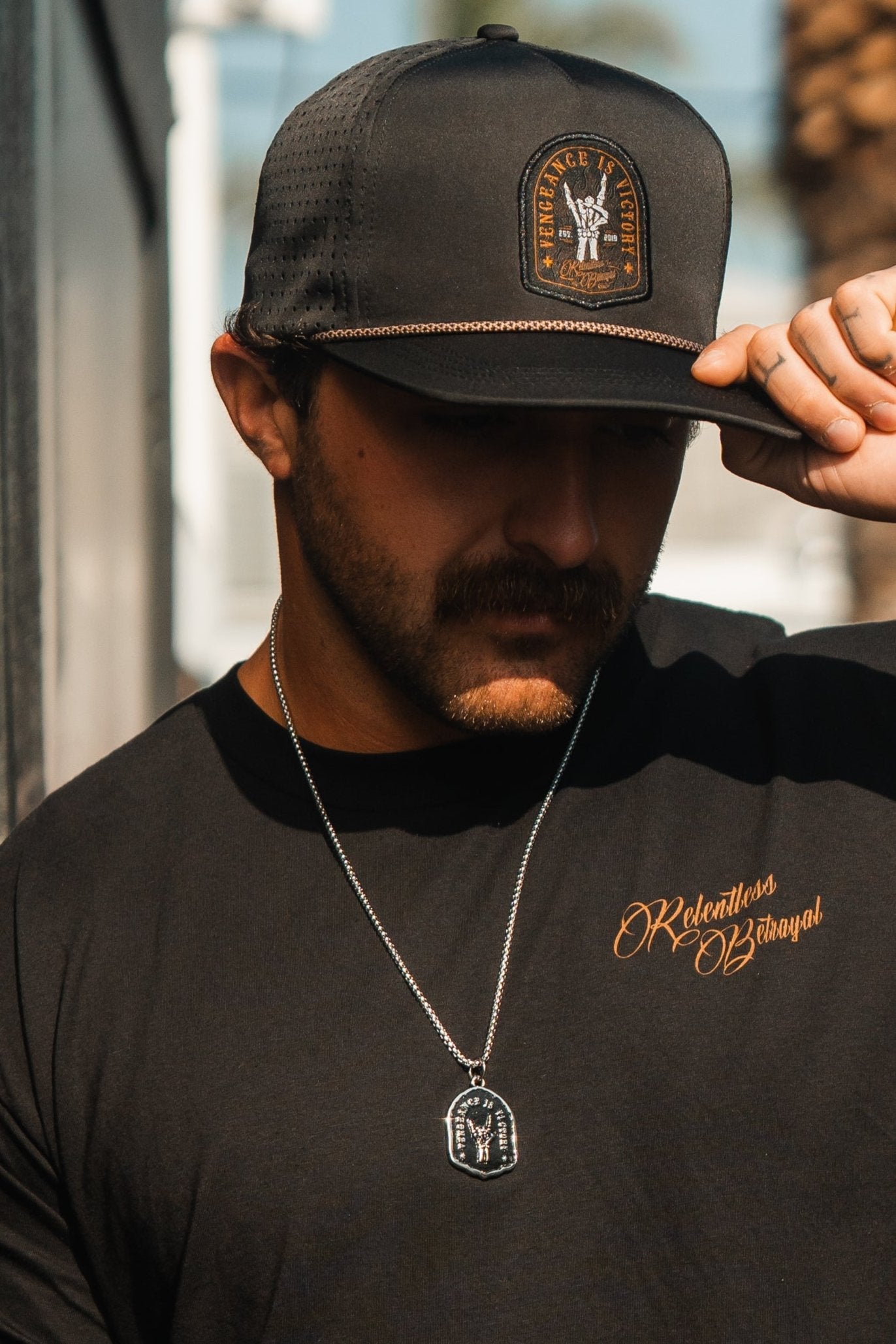 A person outdoors in a Relentless Betrayal Relentless Betrayal t-shirt and black cap adjusts their headwear, while a Vengeance Necklace catches the light, casting shadows that symbolize empowerment and resilience in every contour of their determined stance.