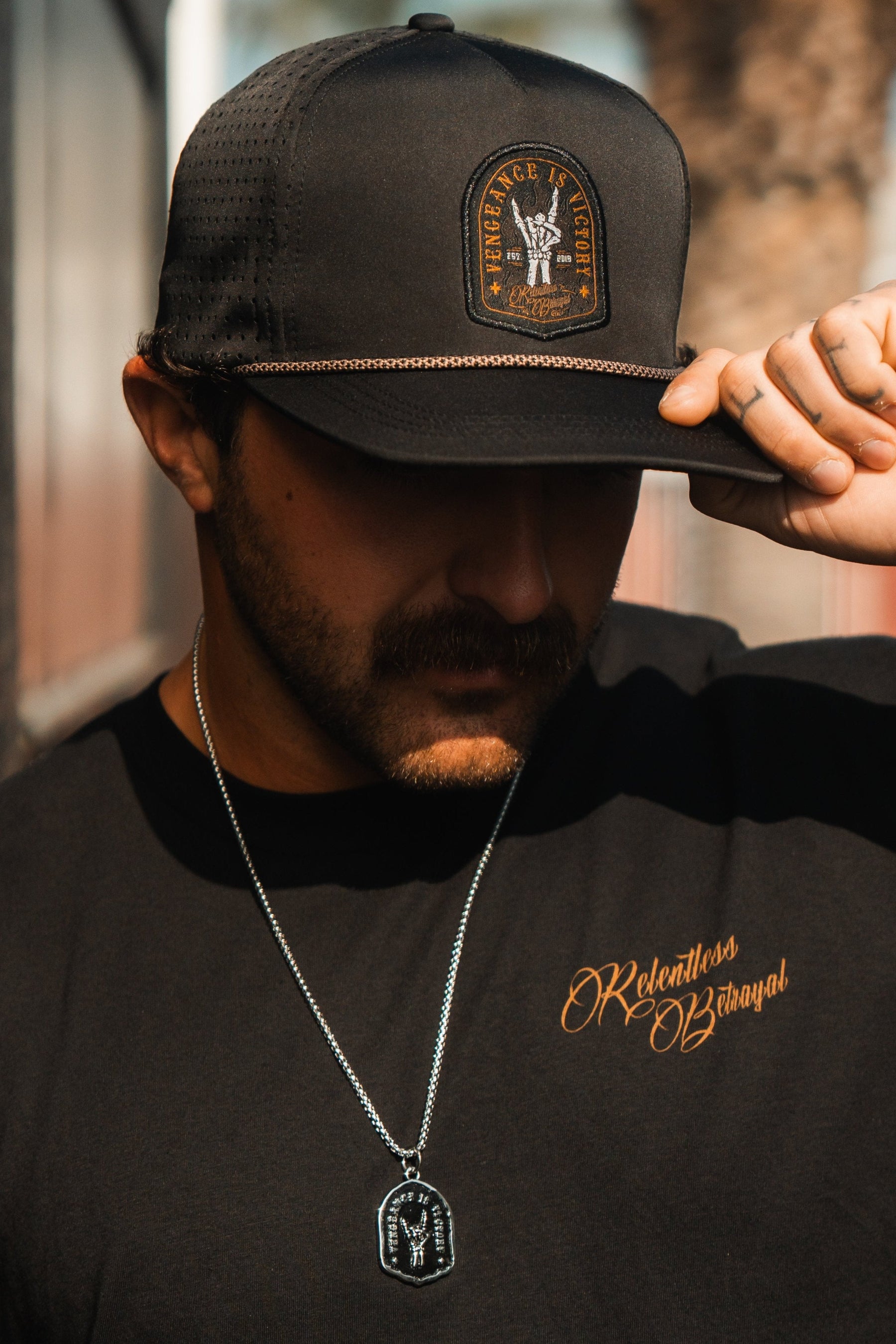 A man in a Relentless Betrayal Vengeance Snapback and black shirt with an embroidered logo, mustached and wearing a pendant necklace, confidently grips the brim. The blurred outdoor background enhances his aura of strength.