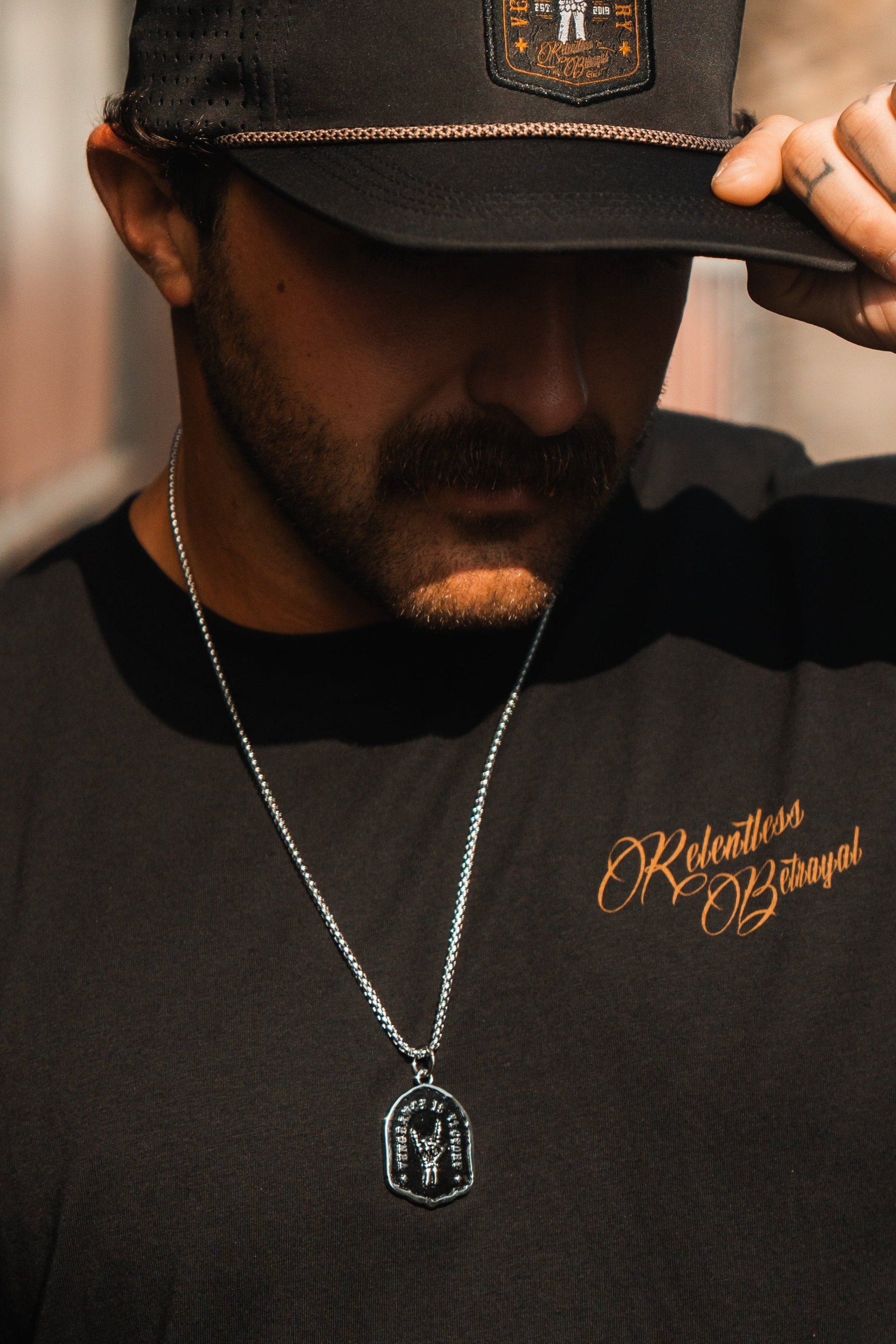 A person in a black cap and Relentless Betrayal black shirt with golden script exudes resilience. Wearing the Vengeance Necklace, symbolizing empowerment, they tilt their head slightly downward against a blurred background.
